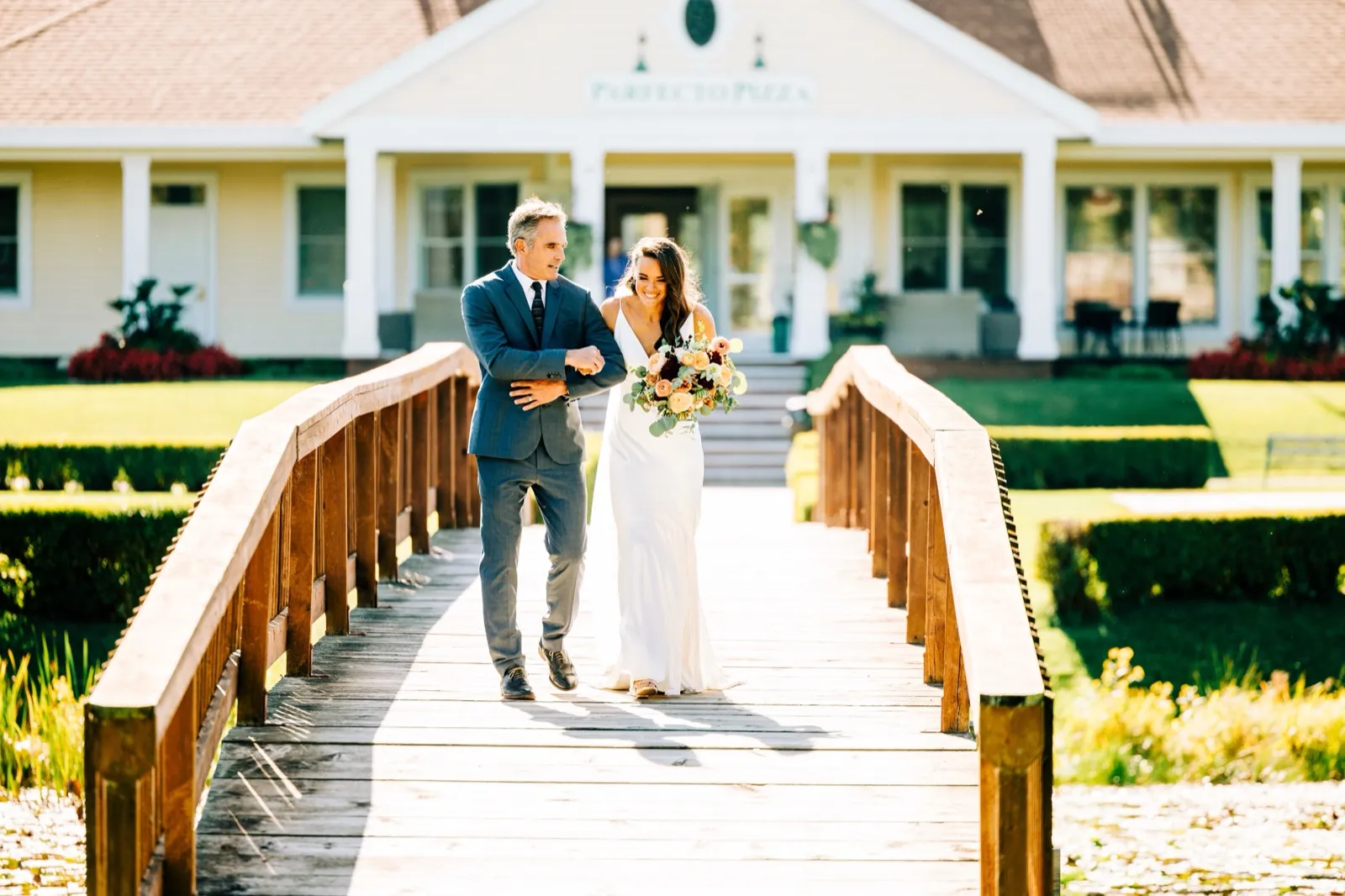 Madison walks arm-in-arm with her father across an arched wooden footbridge toward the ceremony, the lodge behind them at Madden's — Tim Larsen Photography, Brainerd Lakes MN