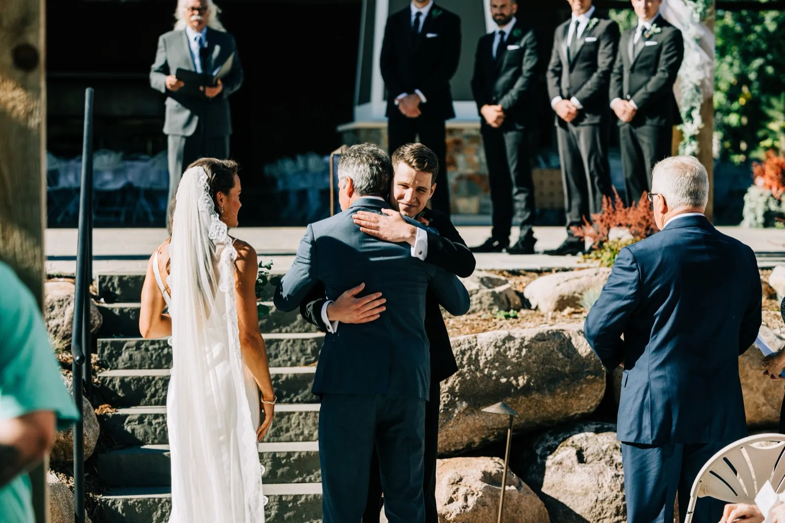 Josh embraces Madison's father in the aisle as she looks on and the groomsmen watch from the stone patio — Tim Larsen Photography, Brainerd Lakes MN