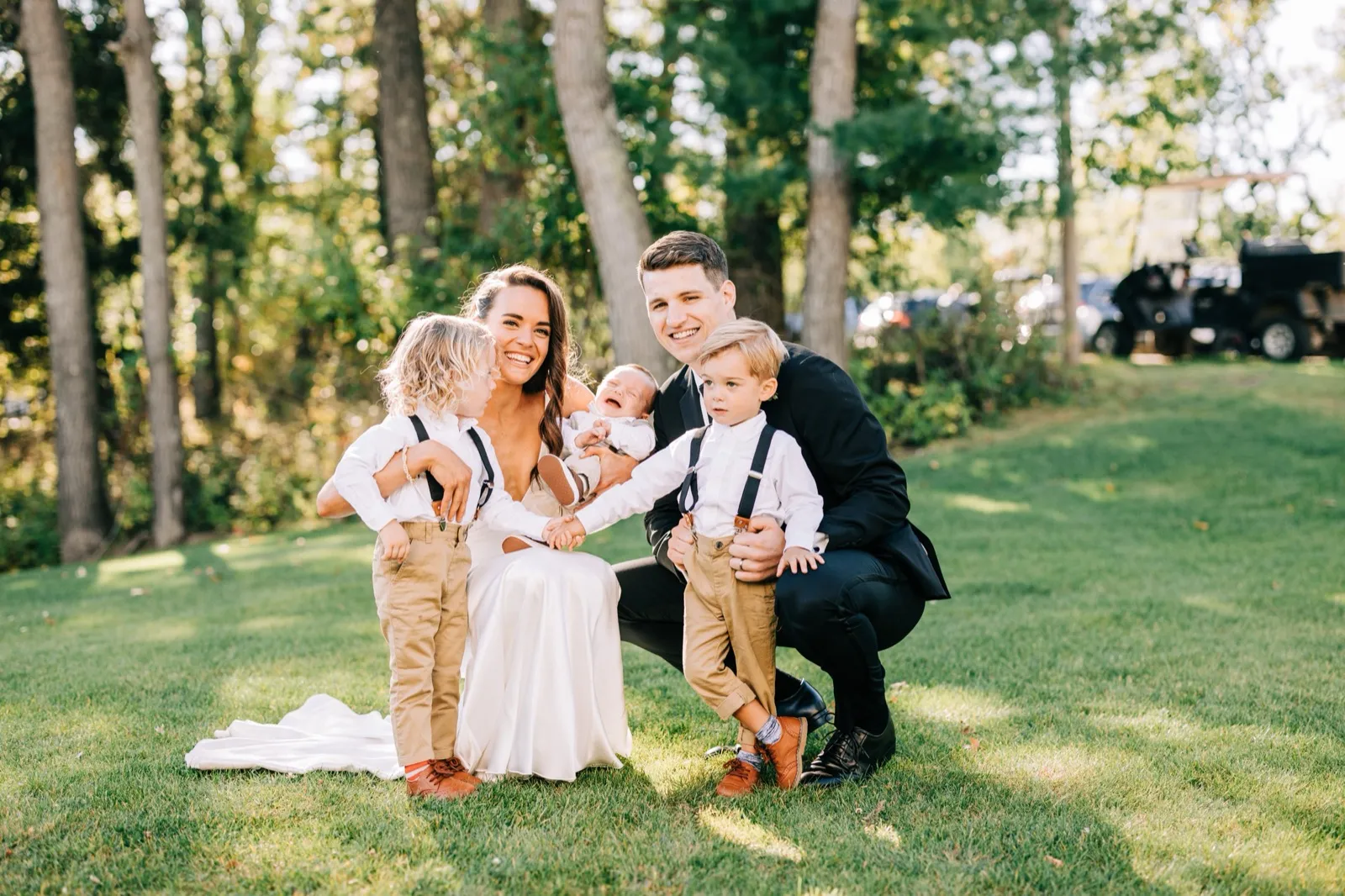 Madison and Josh crouch on a sunlit lawn with two young boys in suspenders and a baby, all laughing together — Tim Larsen Photography, Brainerd Lakes MN