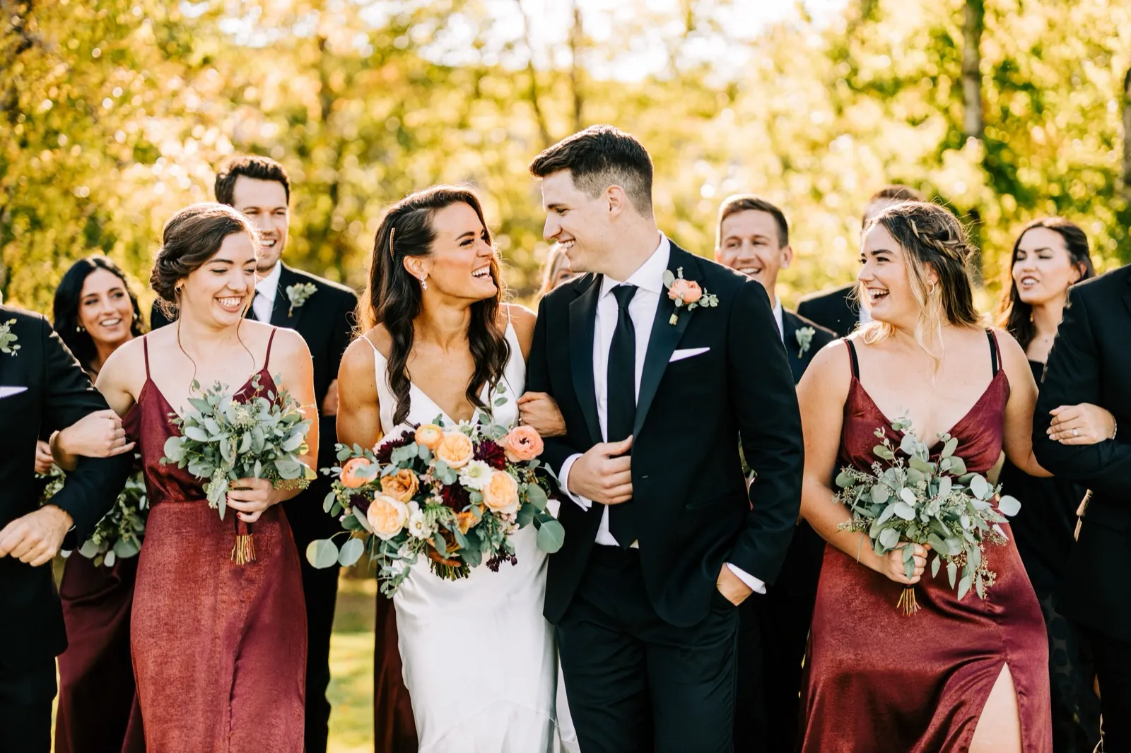 Madison and Josh laugh together as their bridal party walks alongside, all backlit by golden autumn light through the trees — Tim Larsen Photography, Brainerd Lakes MN