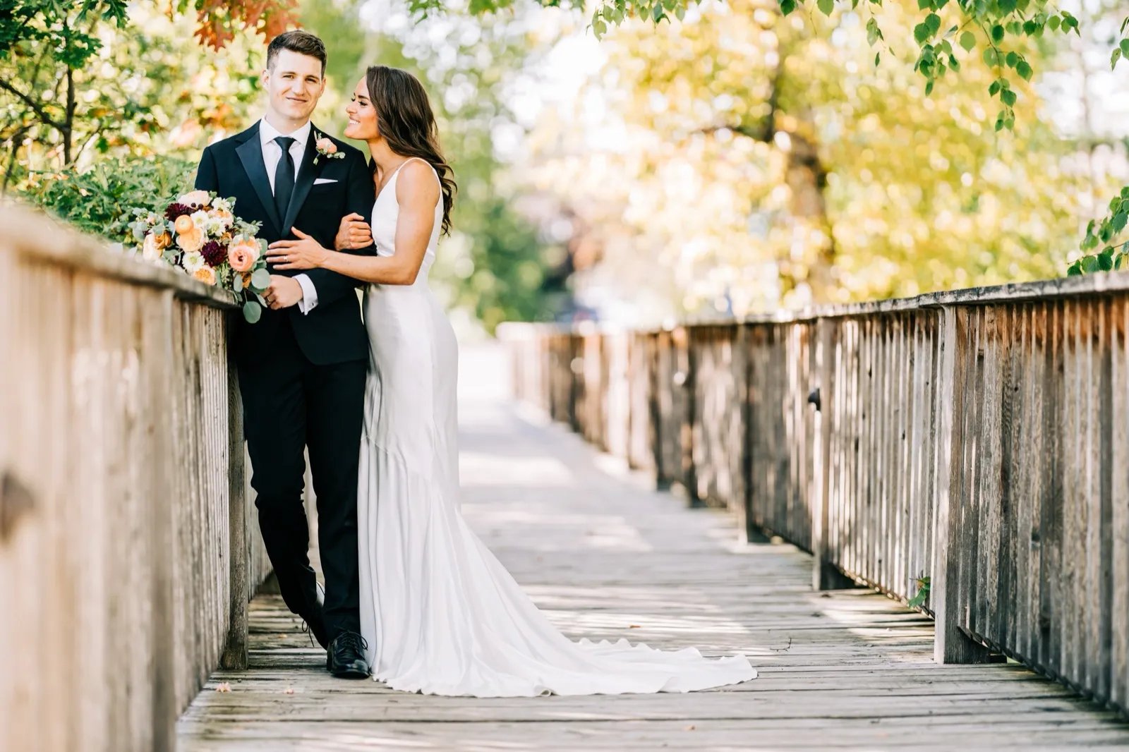 Madison leans into Josh on a wooden footbridge surrounded by autumn foliage, holding a peach-and-burgundy bouquet — Tim Larsen Photography, Brainerd Lakes MN