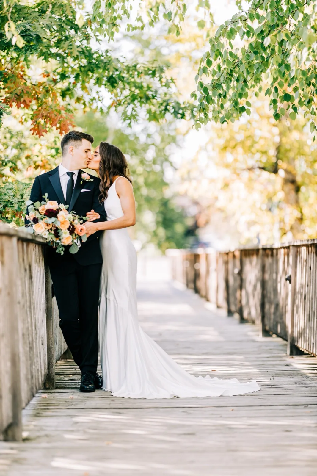 Madison and Josh kiss on a wooden footbridge framed by green and gold leaves, her gown spilling along the planks at Madden's — Tim Larsen Photography, Brainerd Lakes MN