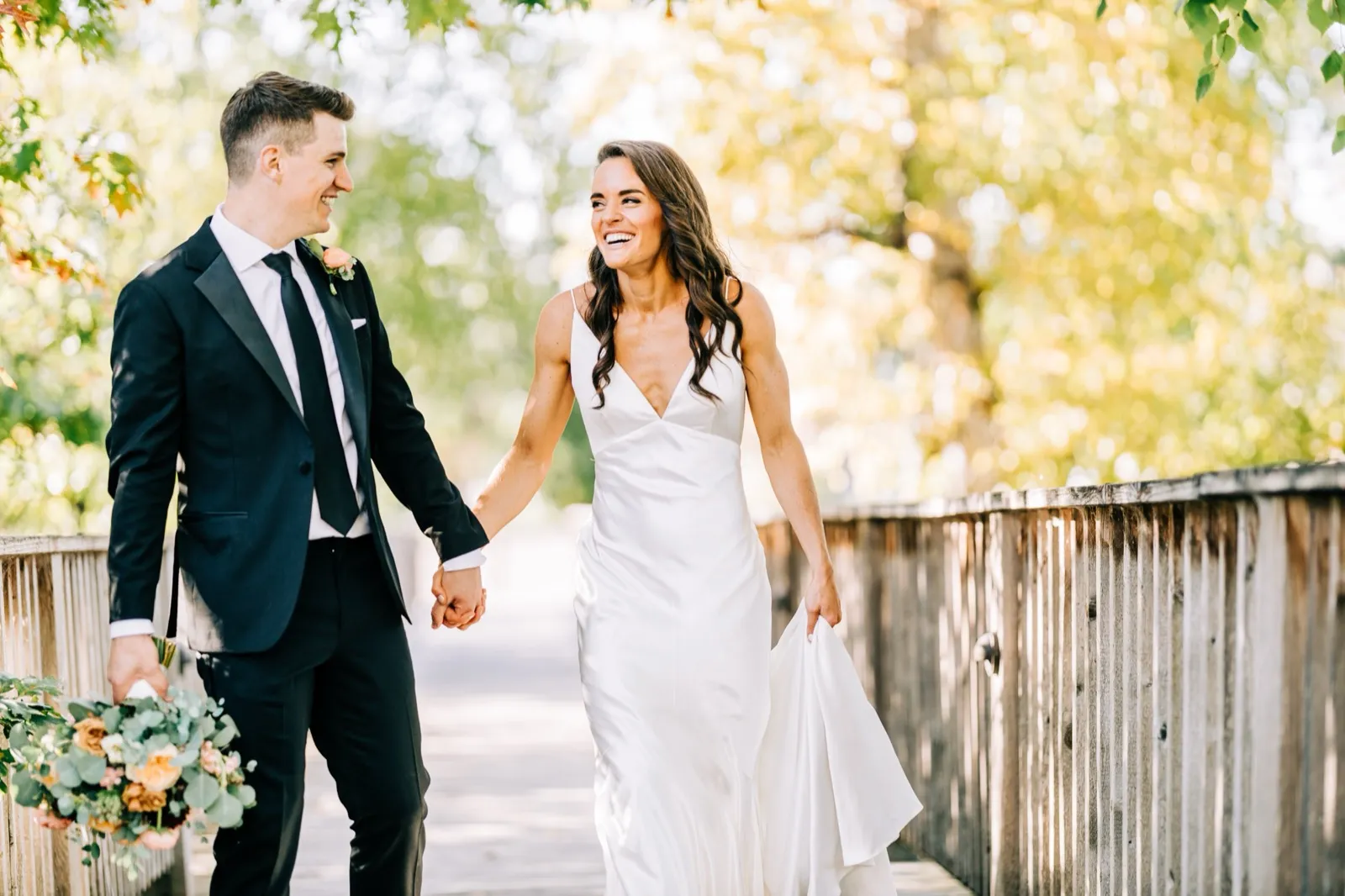 Madison laughs holding Josh's hand as they walk a wooden footbridge framed by sunlit autumn trees — Tim Larsen Photography, Brainerd Lakes MN