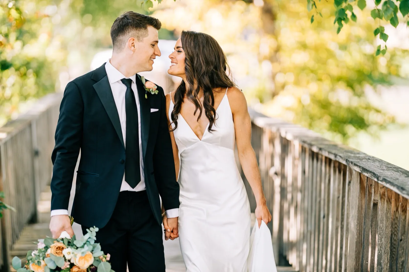 Madison and Josh hold hands and smile at each other walking across a footbridge in backlit autumn foliage — Tim Larsen Photography, Brainerd Lakes MN
