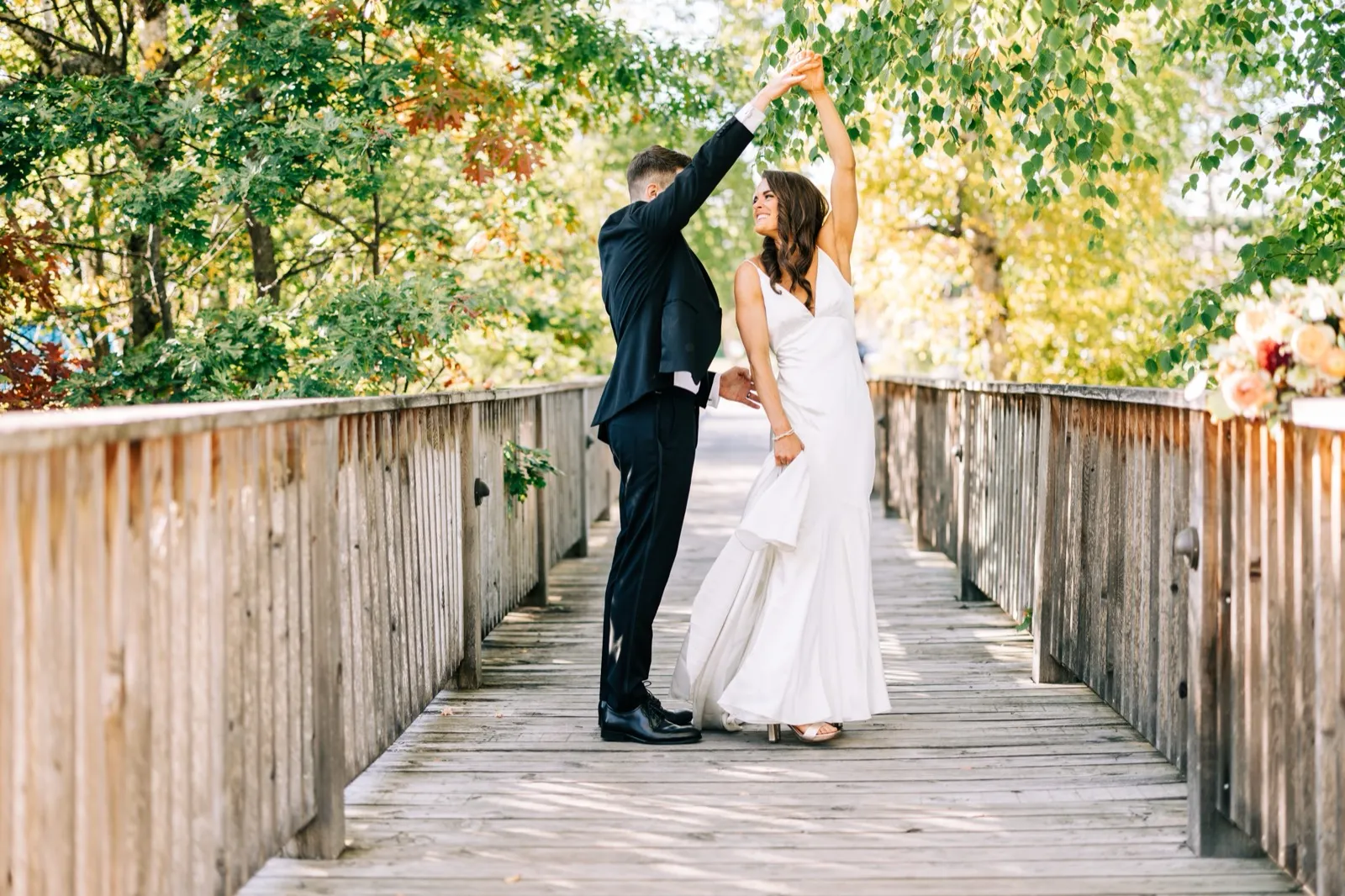 Josh twirls Madison on a wooden footbridge framed by autumn foliage, her dress flaring as she laughs up at him — Tim Larsen Photography, Brainerd Lakes MN