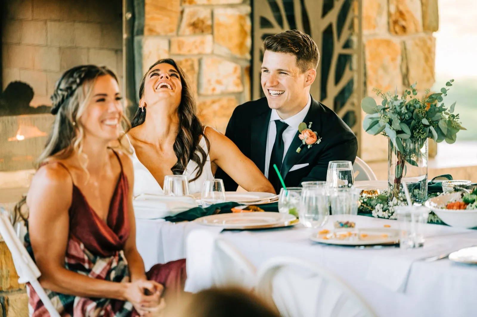 Madison throws her head back laughing between Josh and a bridesmaid at the head table, a stone fireplace behind them at Madden's — Tim Larsen Photography, Brainerd Lakes MN