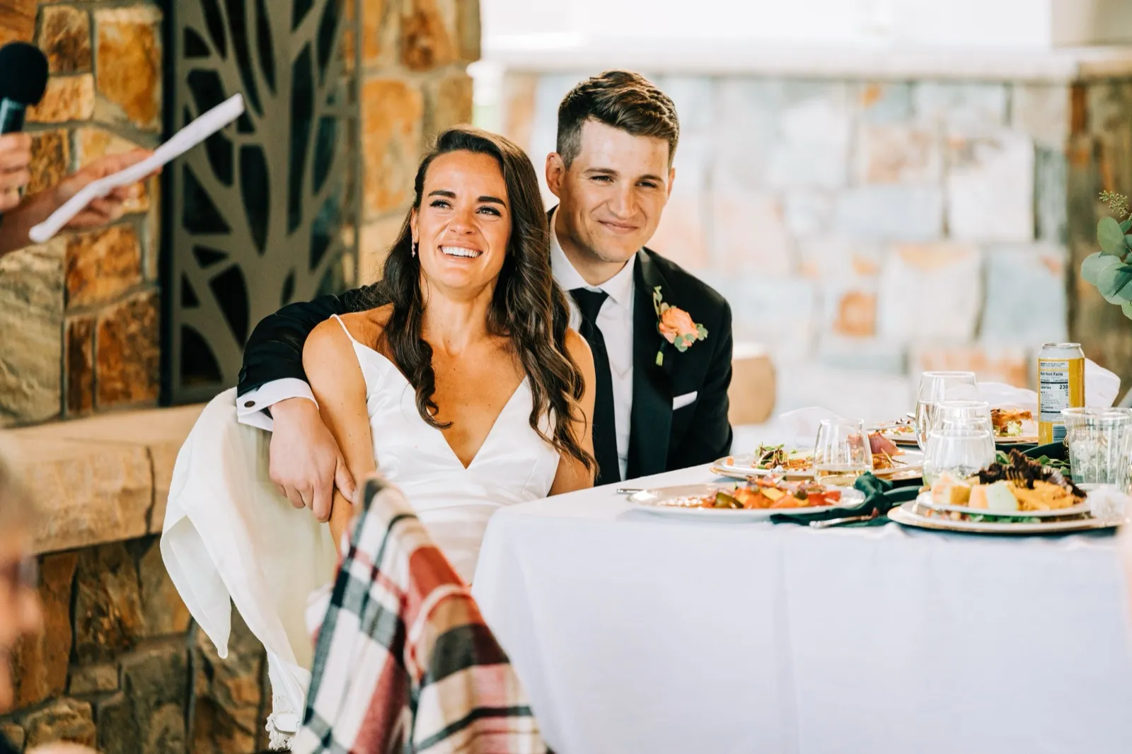 Madison laughs and Josh smiles as they listen to a toast at their sweetheart table by the stone fireplace — Tim Larsen Photography, Brainerd Lakes MN