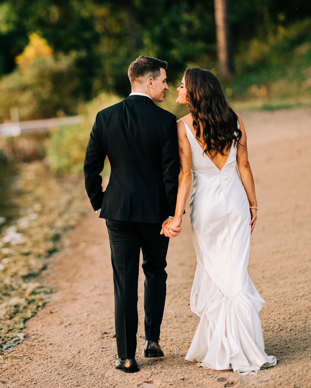 Madison and Josh walk hand in hand along the sandy lakeshore at golden hour, turning to each other with the dock and Gull Lake behind — Tim Larsen Photography, Brainerd Lakes MN