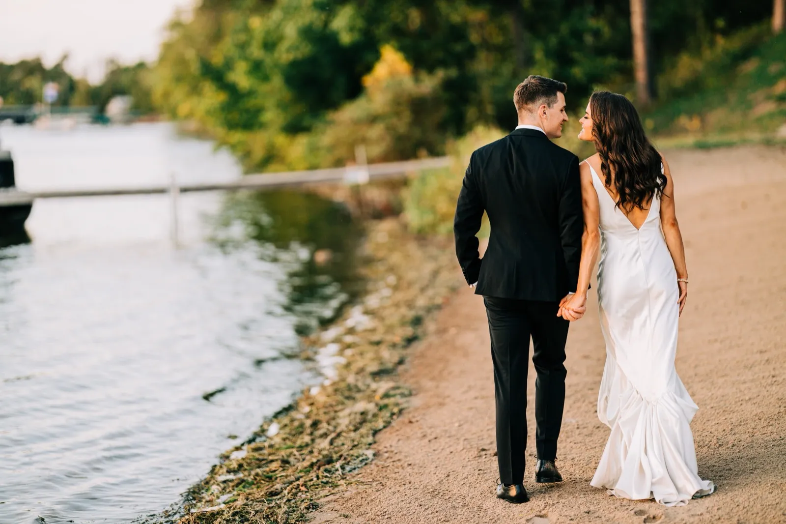 Madison and Josh walk hand in hand along the sandy lakeshore at golden hour, turning to each other with the dock and Gull Lake behind — Tim Larsen Photography, Brainerd Lakes MN