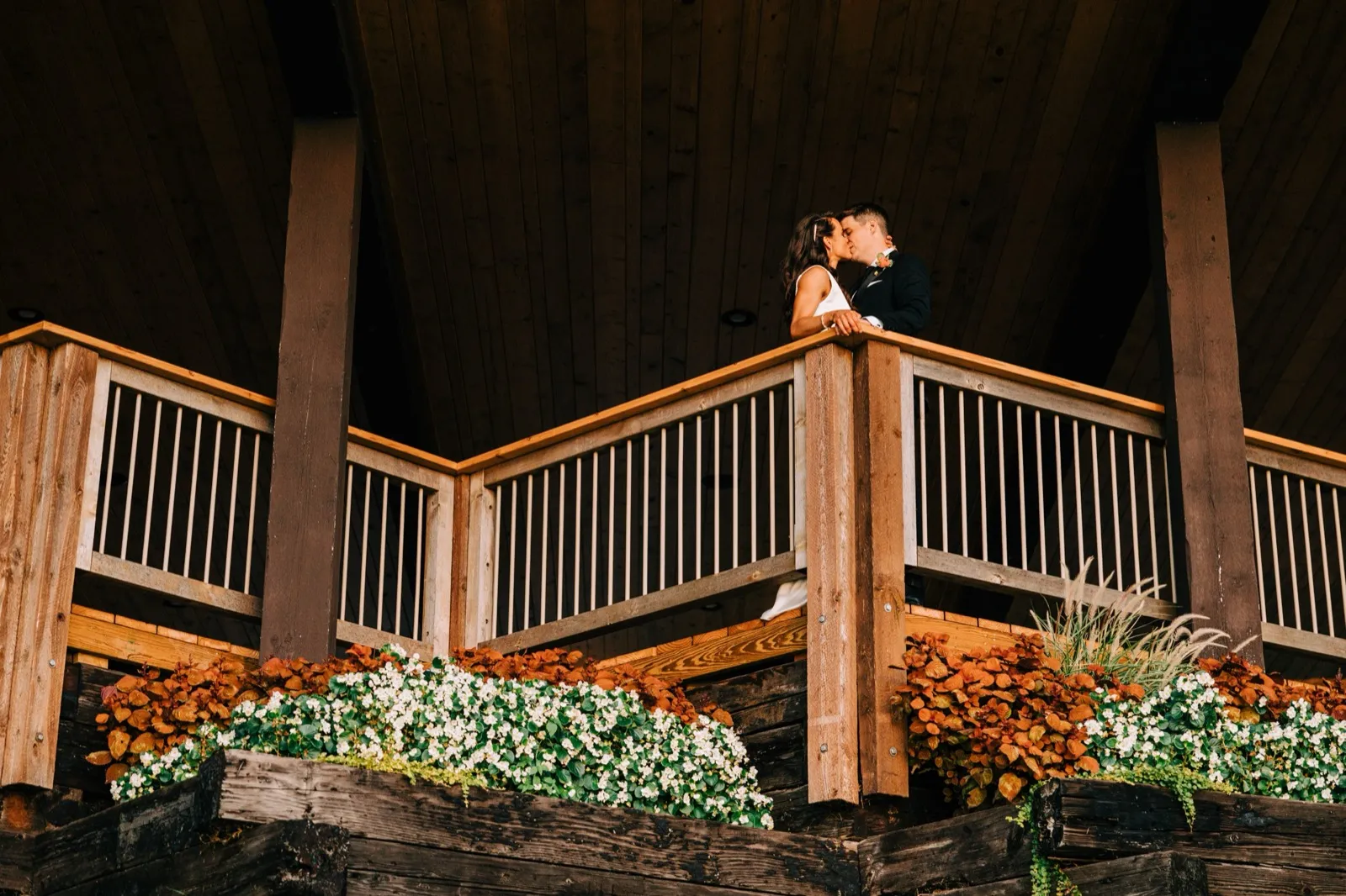 Madison and Josh kiss on a timber lodge balcony, planter boxes of white flowers and autumn foliage spilling below — Tim Larsen Photography, Brainerd Lakes MN