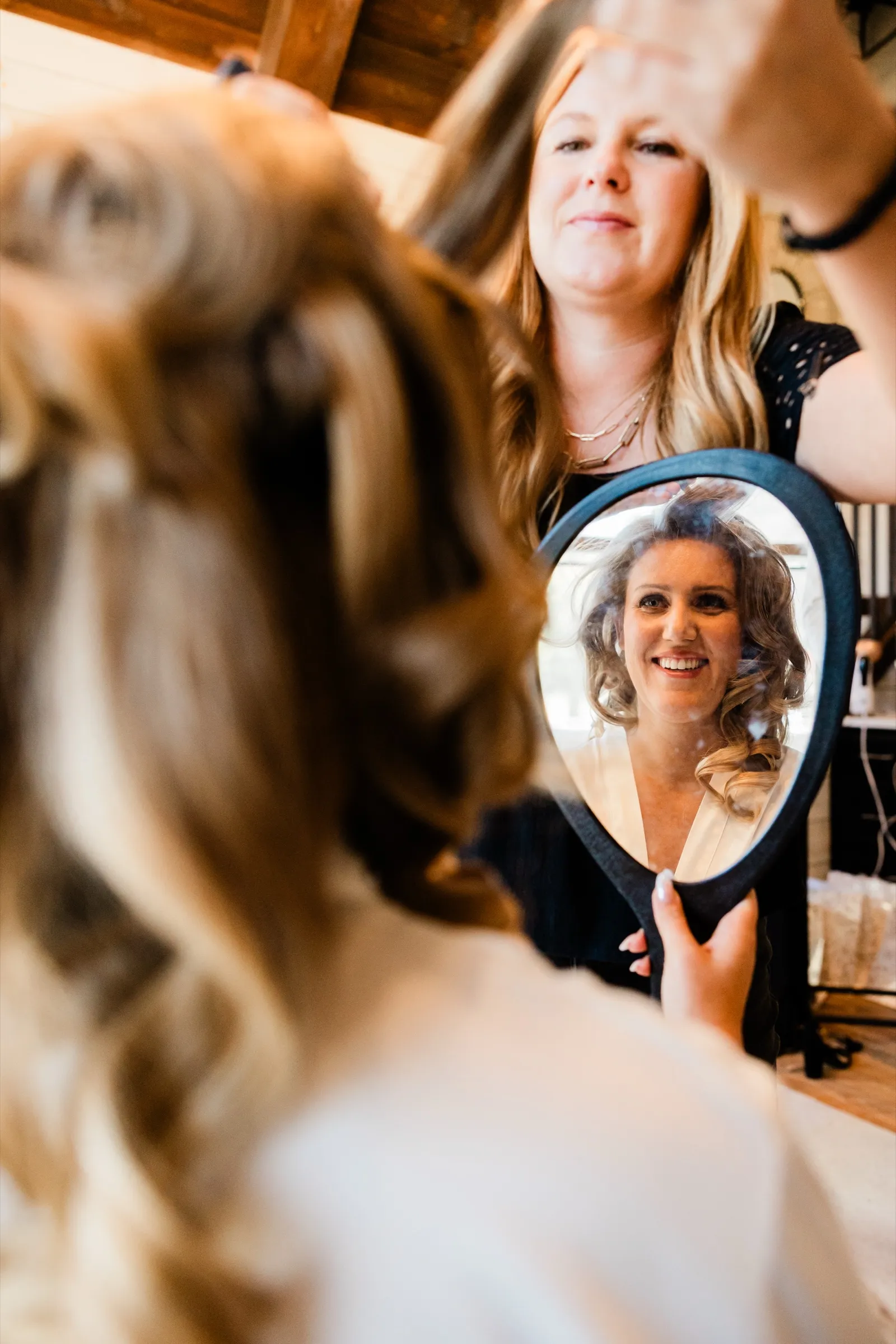 A bride watches her own reflection in a handheld mirror as a stylist finishes her hair, the moment caught over her shoulder without her noticing the camera — Tim Larsen Photography, Brainerd Lakes MN