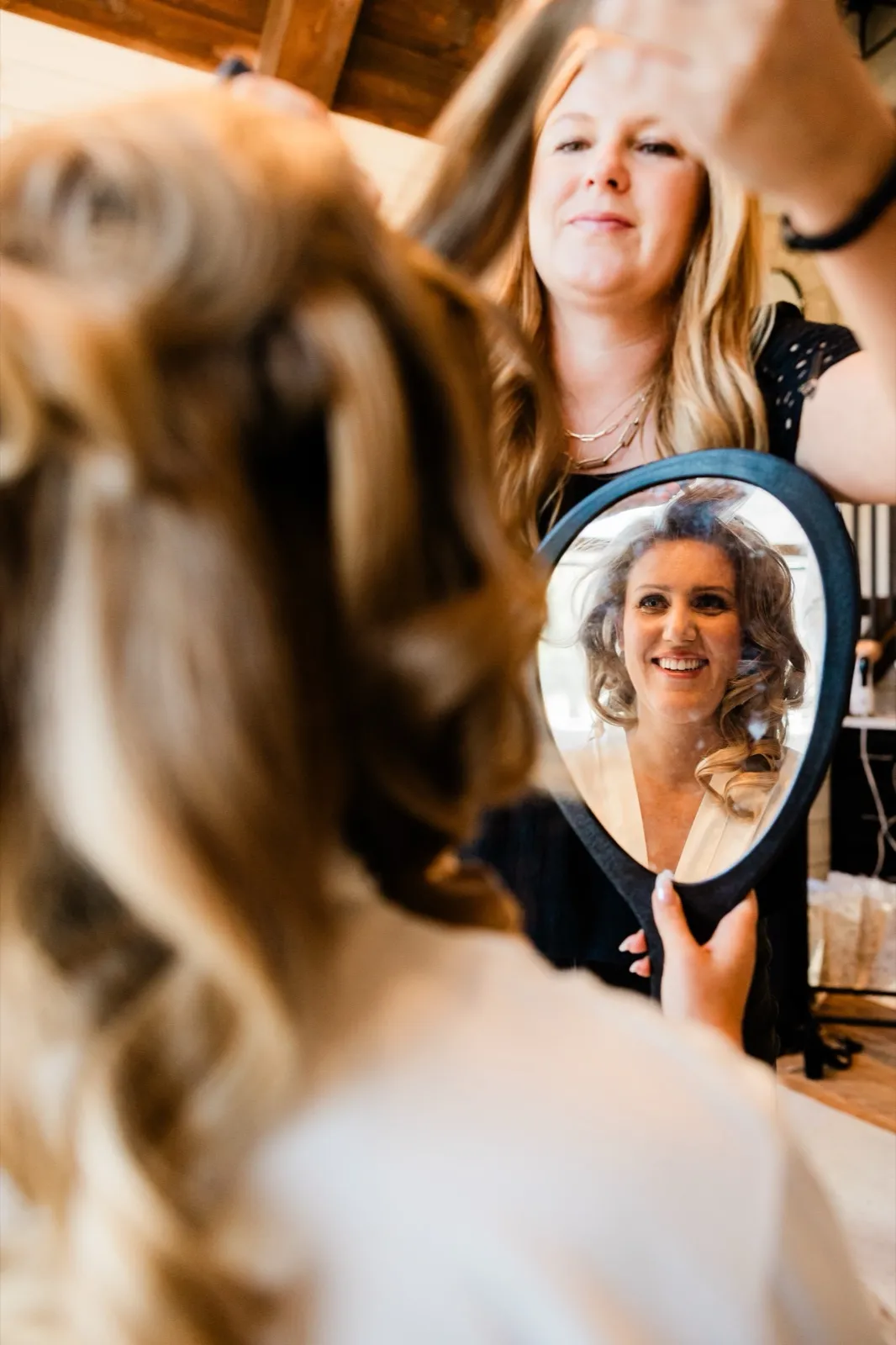 A bride watches her own reflection in a handheld mirror as a stylist finishes her hair, the moment caught over her shoulder without her noticing the camera — Tim Larsen Photography, Brainerd Lakes MN