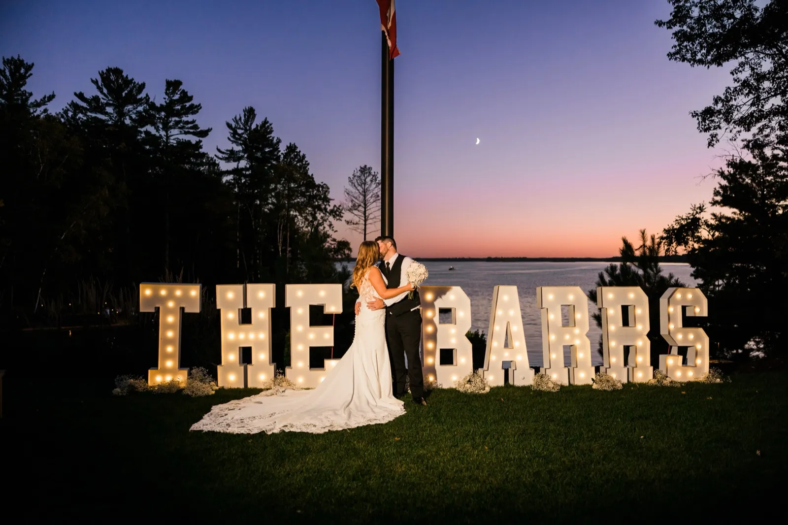 A couple kisses in front of glowing marquee letters reading 'THE BARRS' at dusk, a crescent moon and pink sunset over the lake behind them — the light you get when the timeline is planned backward from sunset — Tim Larsen Photography, Brainerd Lakes MN