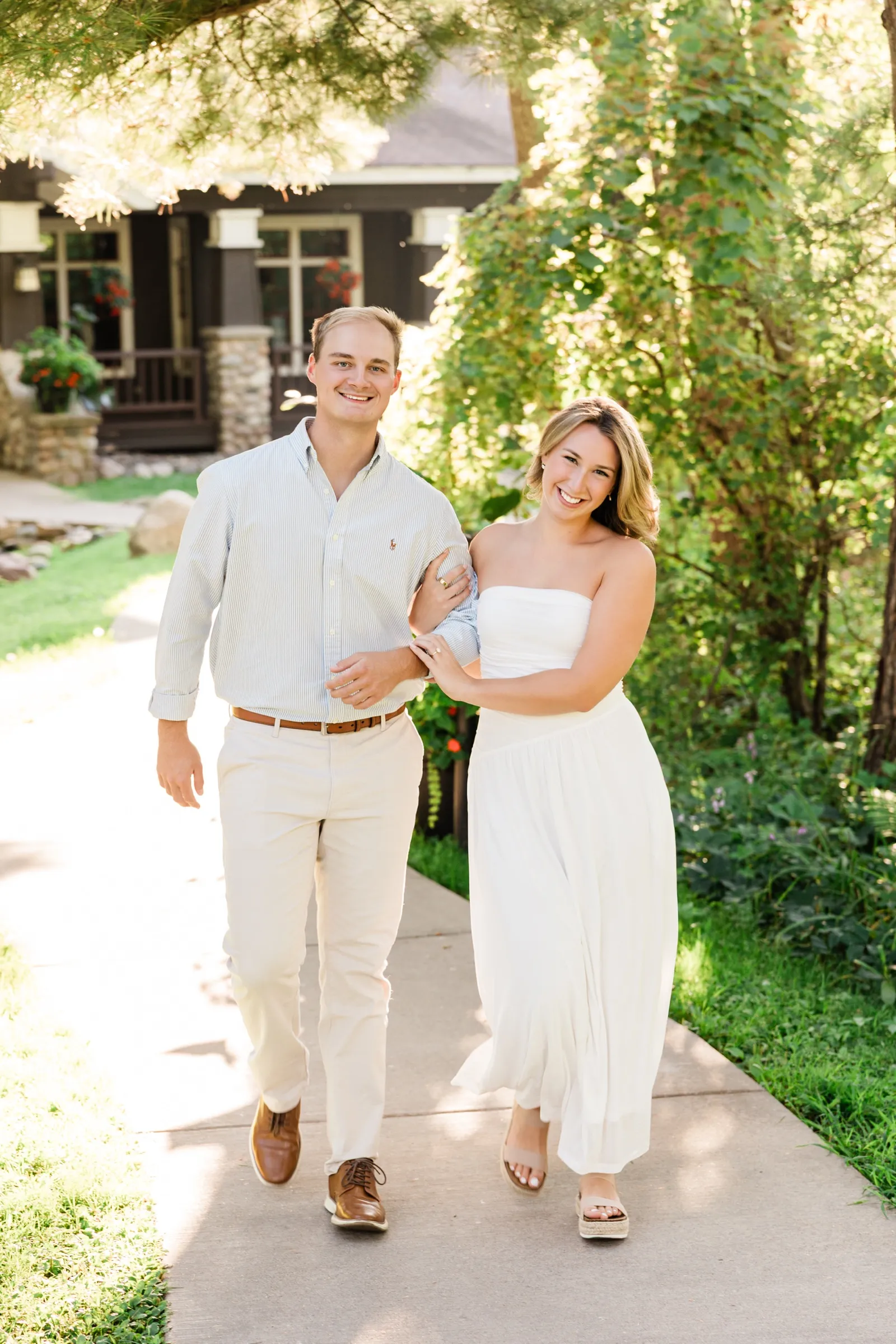 Olivia hugs Adam's arm as they walk a sunlit sidewalk past a craftsman-style building at Grand View Lodge — Tim Larsen Photography