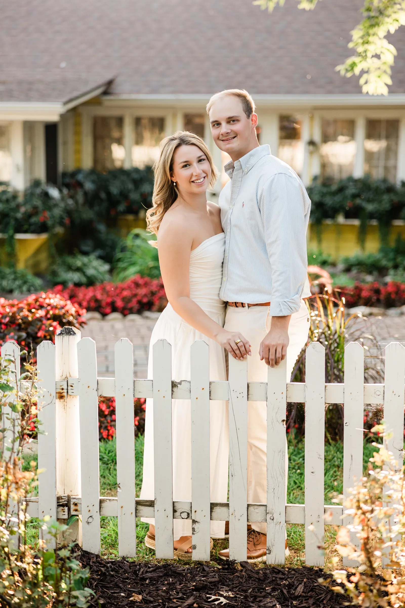 Olivia and Adam stand behind a white picket gate with the yellow Italian Gardens restaurant and red flower beds behind them at Grand View Lodge — Tim Larsen Photography