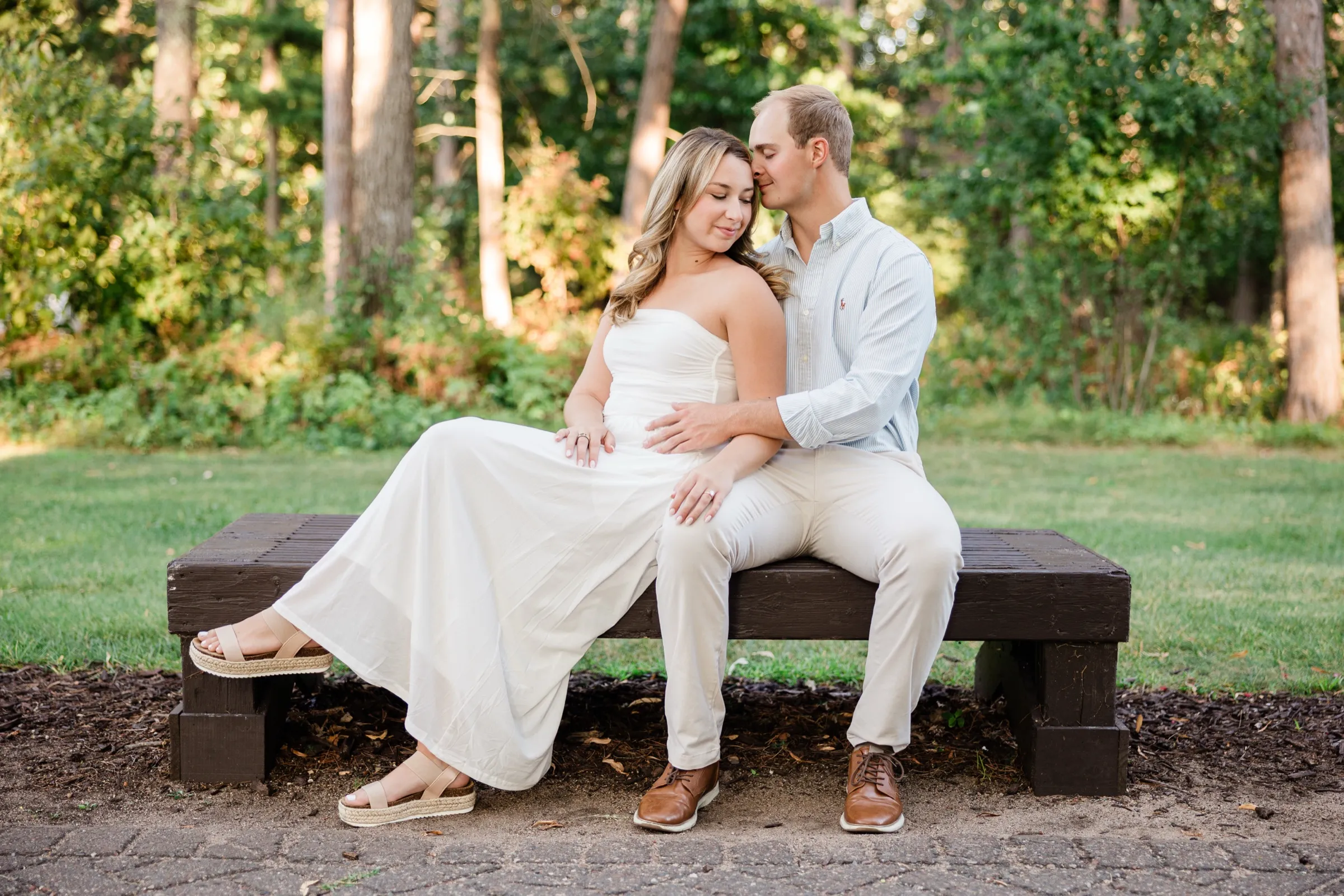 Olivia and Adam sit close on a wooden bench at the pine forest edge, Adam kissing her temple — Tim Larsen Photography