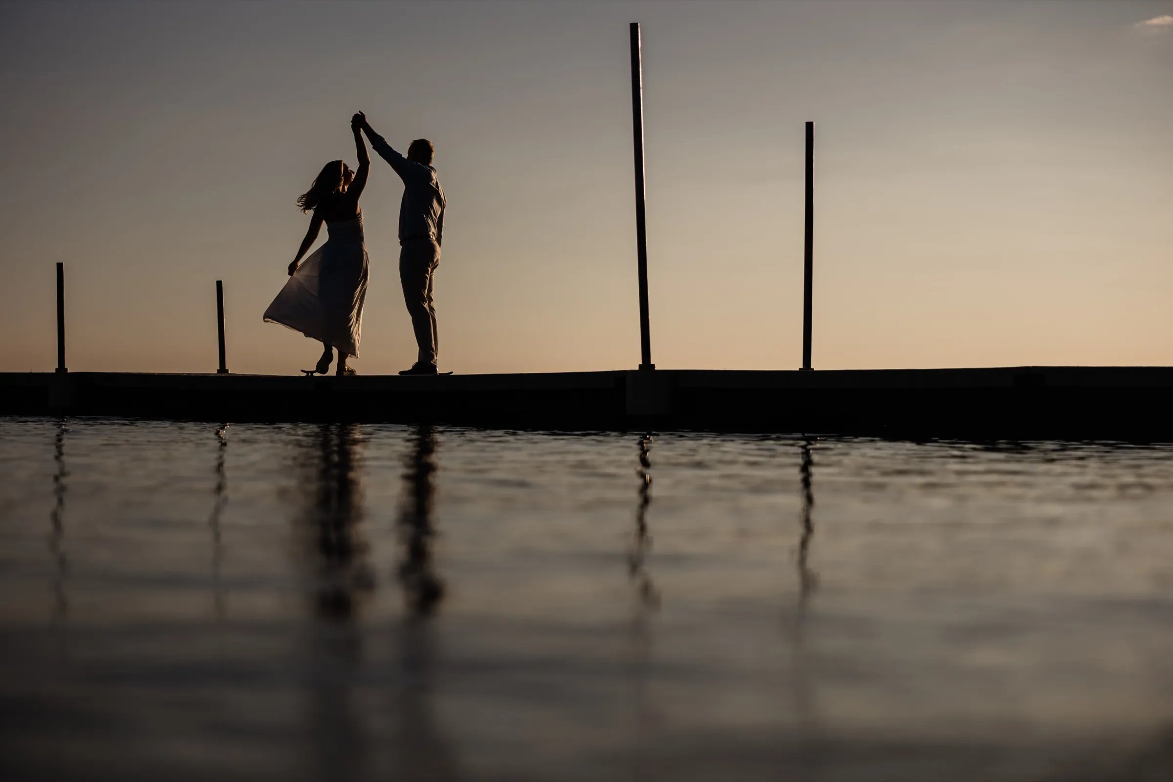 Adam twirls Olivia in silhouette on the dock at dusk, her dress flaring above the water's reflection — Tim Larsen Photography