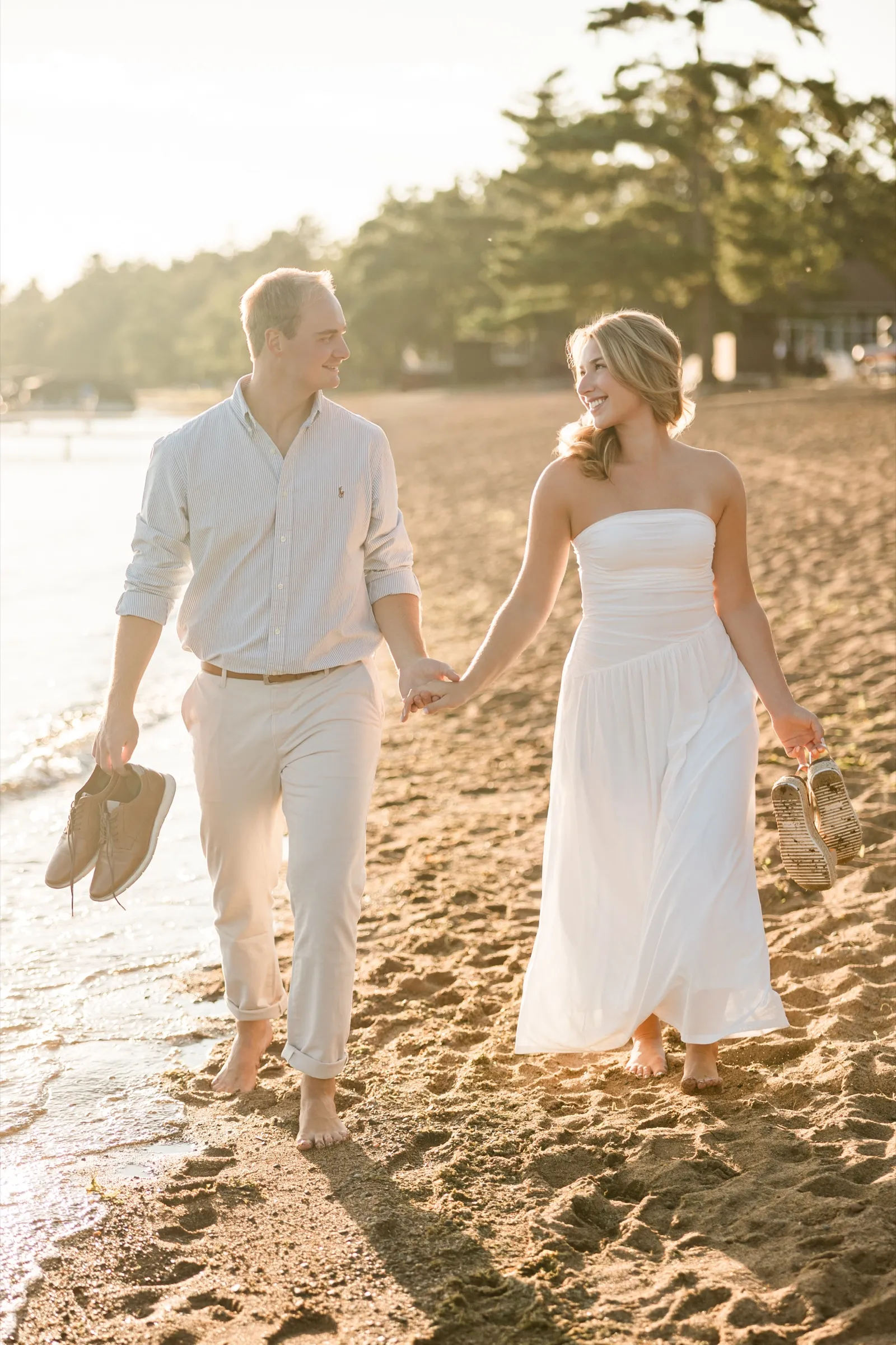 Olivia and Adam walk barefoot down the golden sand beach at sunset, carrying their shoes — Tim Larsen Photography