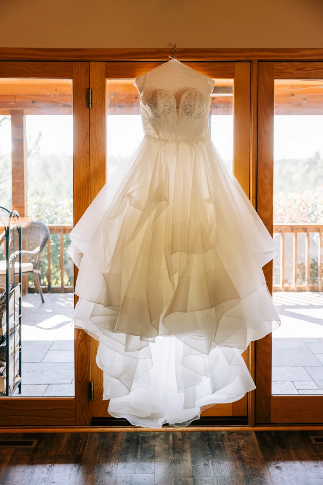 A wedding gown with a lace bodice and layered tulle skirt hangs from french doors, backlit by morning sun spilling onto a wood floor — Tim Larsen Photography, Brainerd Lakes MN