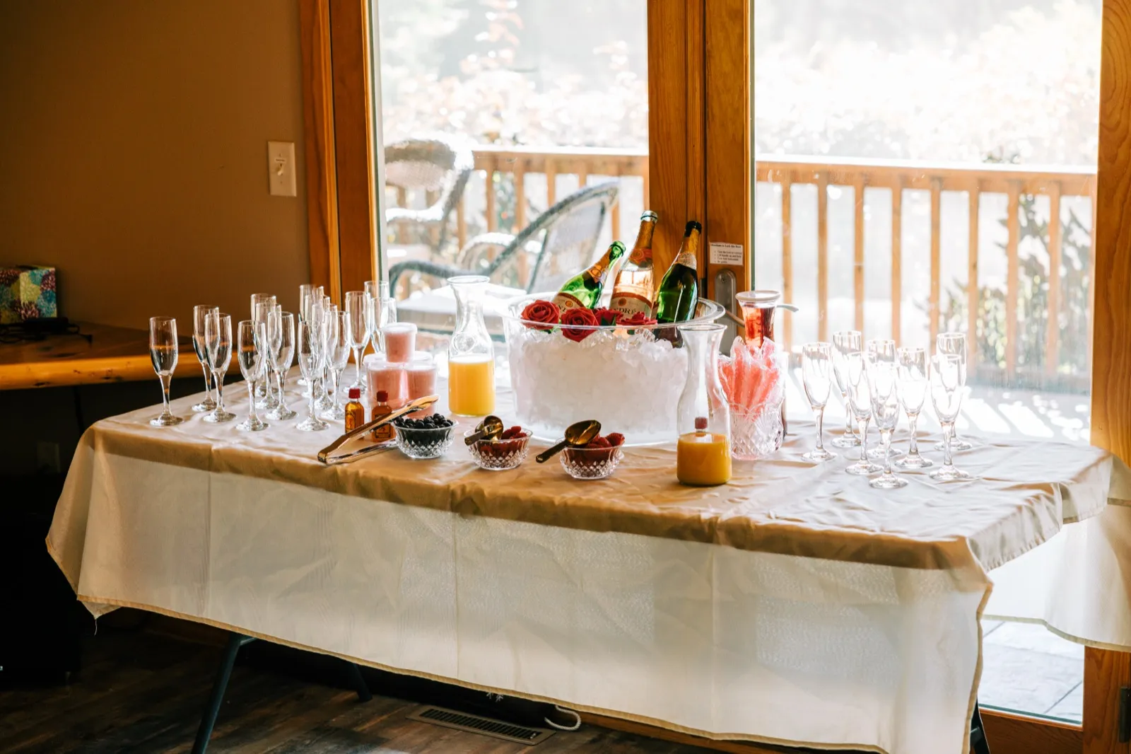 A mimosa bar with champagne flutes, bottles on ice, juice carafes, and bowls of berries arranged on a draped table in front of a sunlit window — Tim Larsen Photography, Brainerd Lakes MN