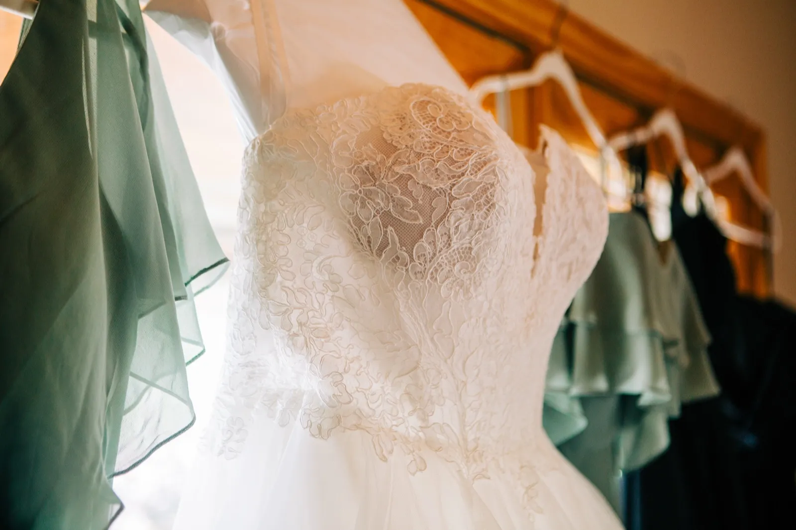 A lace wedding gown hangs in window light beside a row of sage green bridesmaid dresses on wooden hangers — Tim Larsen Photography, Brainerd Lakes MN