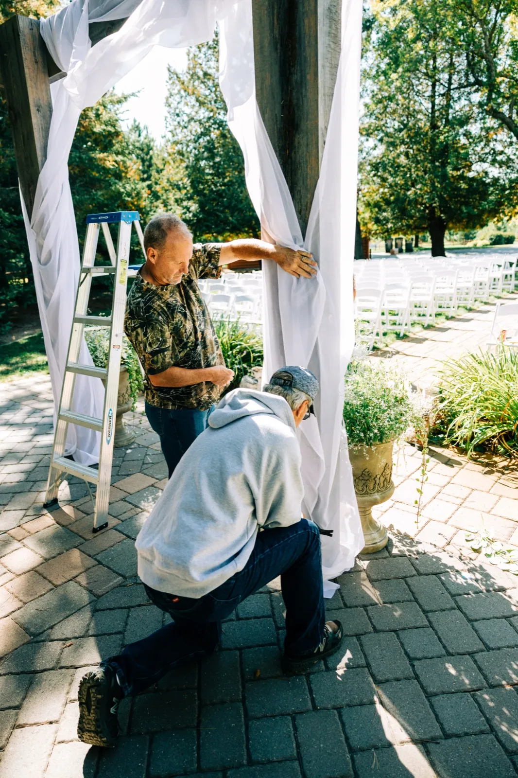 Two men in casual clothes assemble white fabric drapery on a wooden ceremony arch beside a stepladder, with rows of white chairs visible in the background. — Tim Larsen Photography, Brainerd Lakes MN