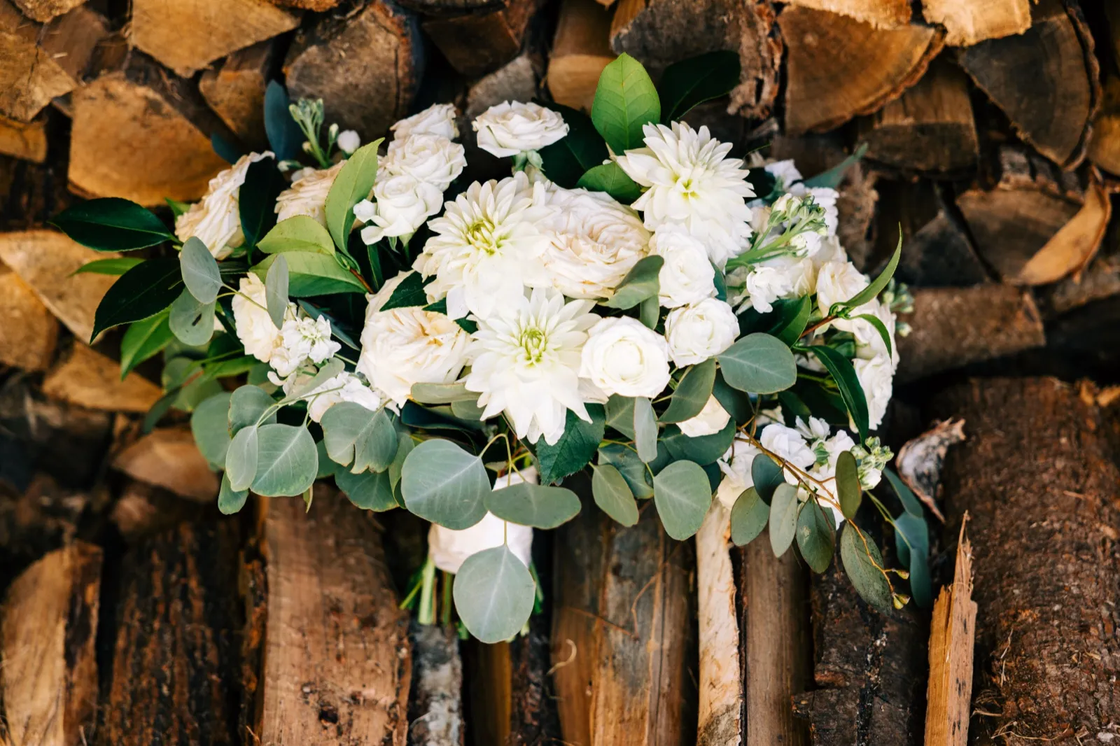 An ivory bridal bouquet of dahlias, roses, and eucalyptus rests against a stacked woodpile backdrop — Tim Larsen Photography, Brainerd Lakes MN
