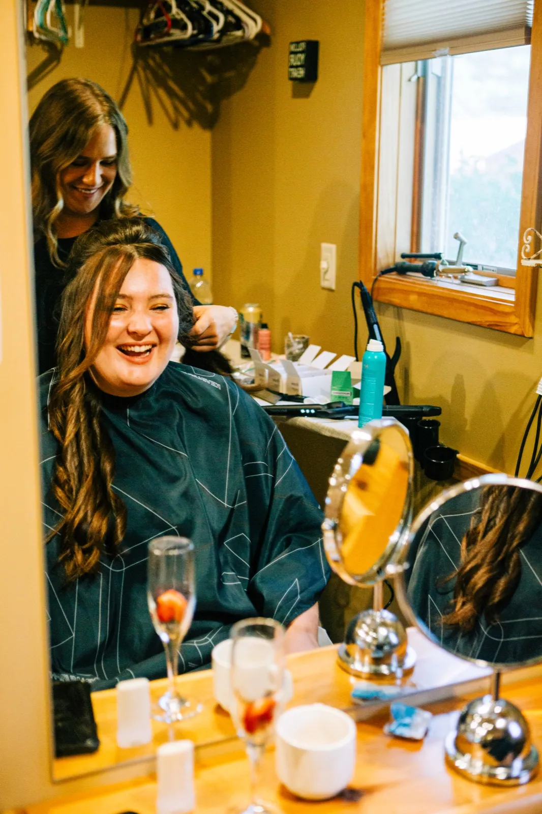 The bride laughs with her eyes closed as a stylist works on her hair, seen in the reflection of a vanity mirror surrounded by champagne flutes and styling products — Tim Larsen Photography, Brainerd Lakes MN