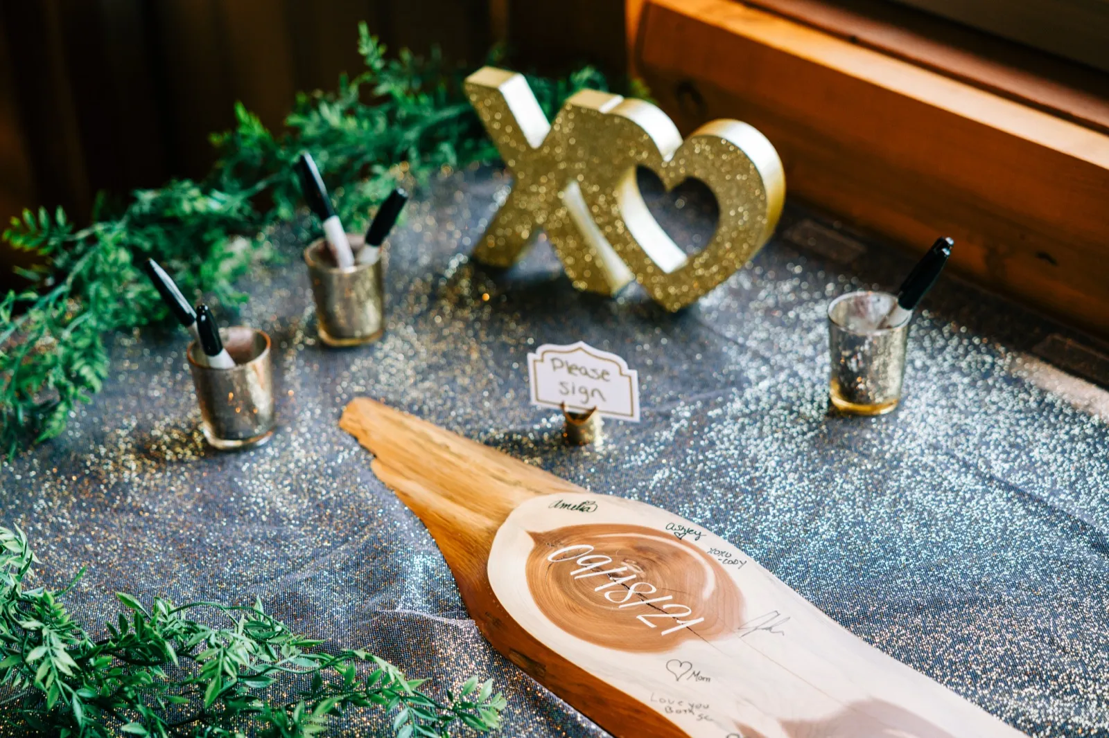 A wood-slab guest book signed with the wedding date 09/18/21 sits on a sequined table beside a gold XO sign, greenery, and mercury glass cups holding pens — Tim Larsen Photography, Brainerd Lakes MN
