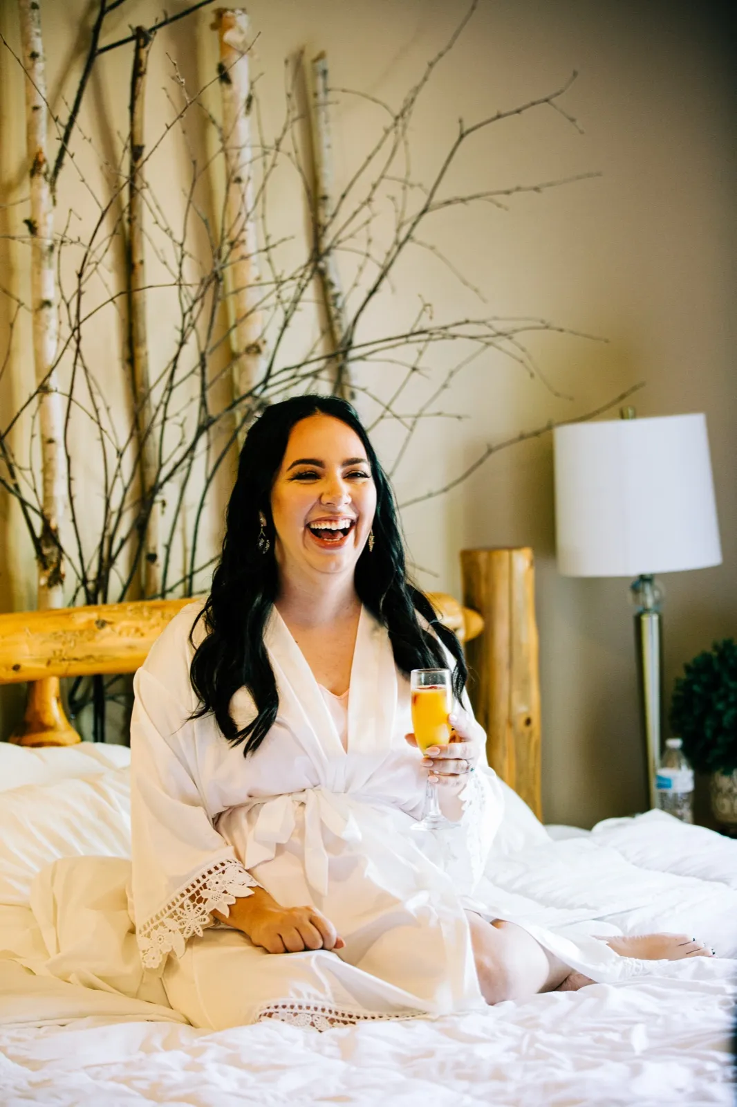 Taylor sits on a bed in a white lace robe holding a mimosa, laughing openly with birch branches arranged behind her. — Tim Larsen Photography, Brainerd Lakes MN