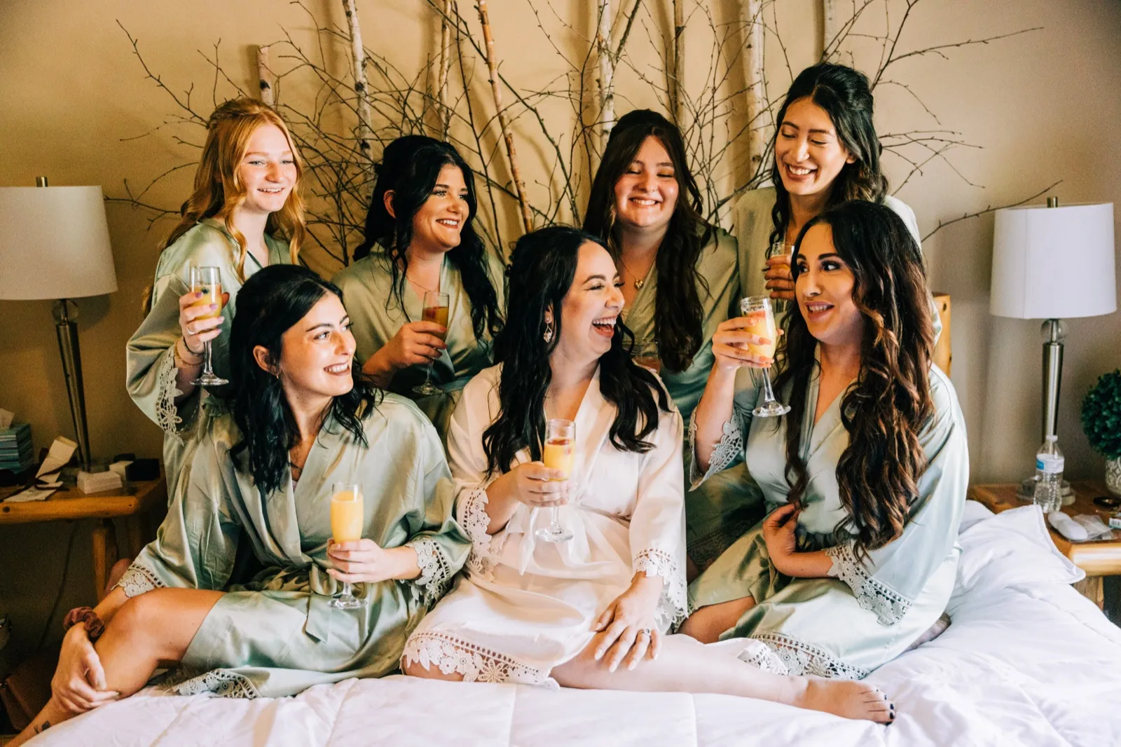 The bride laughs with her six bridesmaids in matching sage robes while raising mimosa glasses on a bed in front of a birch branch backdrop — Tim Larsen Photography, Brainerd Lakes MN