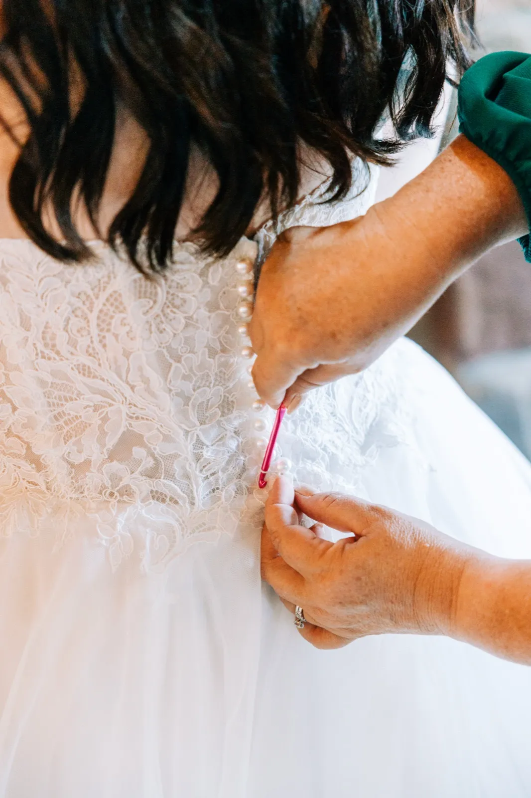 A helper uses a small crochet hook to fasten the row of pearl buttons down the back of the bride's lace wedding dress — Tim Larsen Photography, Brainerd Lakes MN