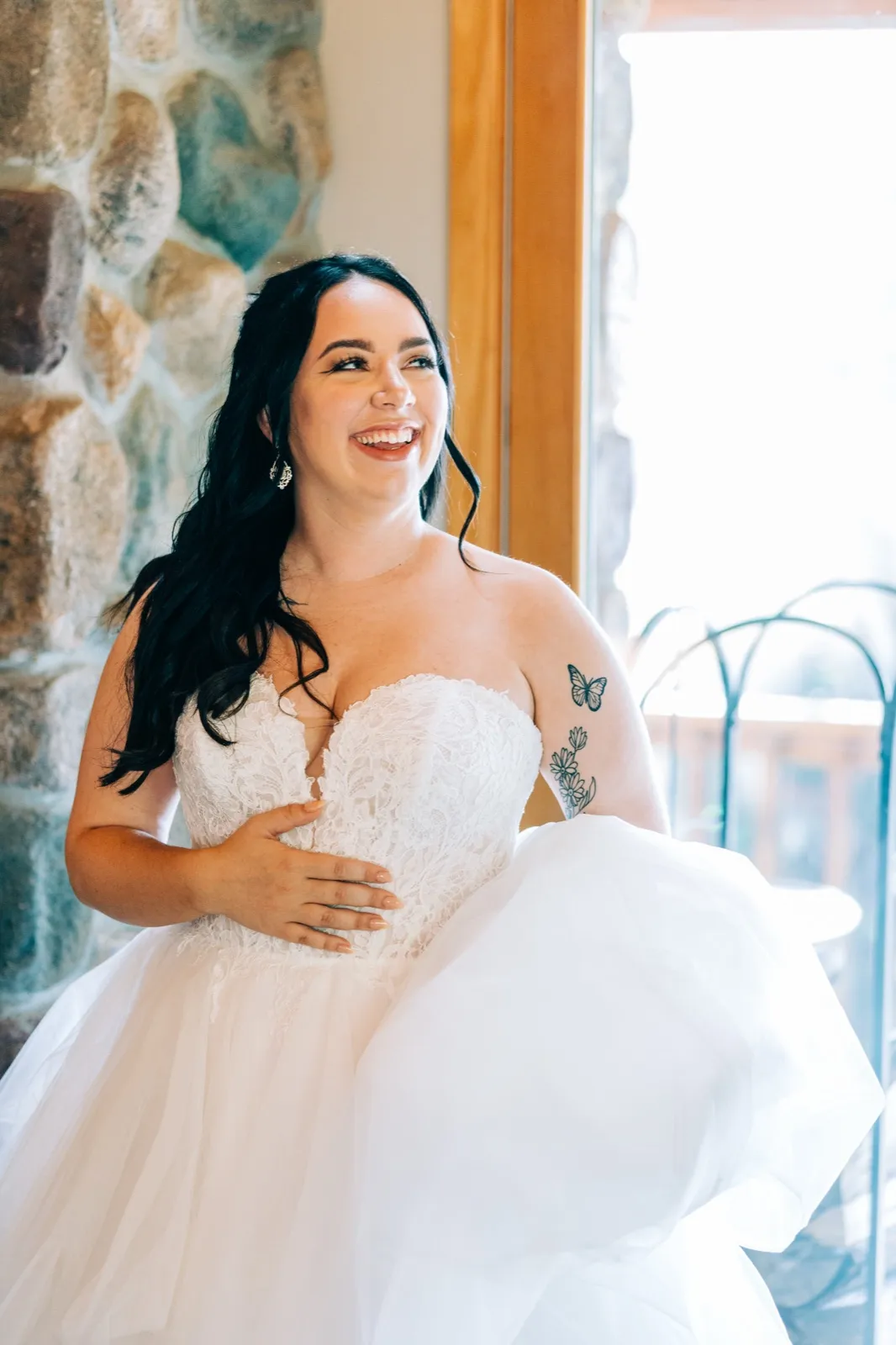 Taylor laughs while gathering her tulle skirt, standing in window light beside a stone fireplace in the bridal suite. — Tim Larsen Photography, Brainerd Lakes MN