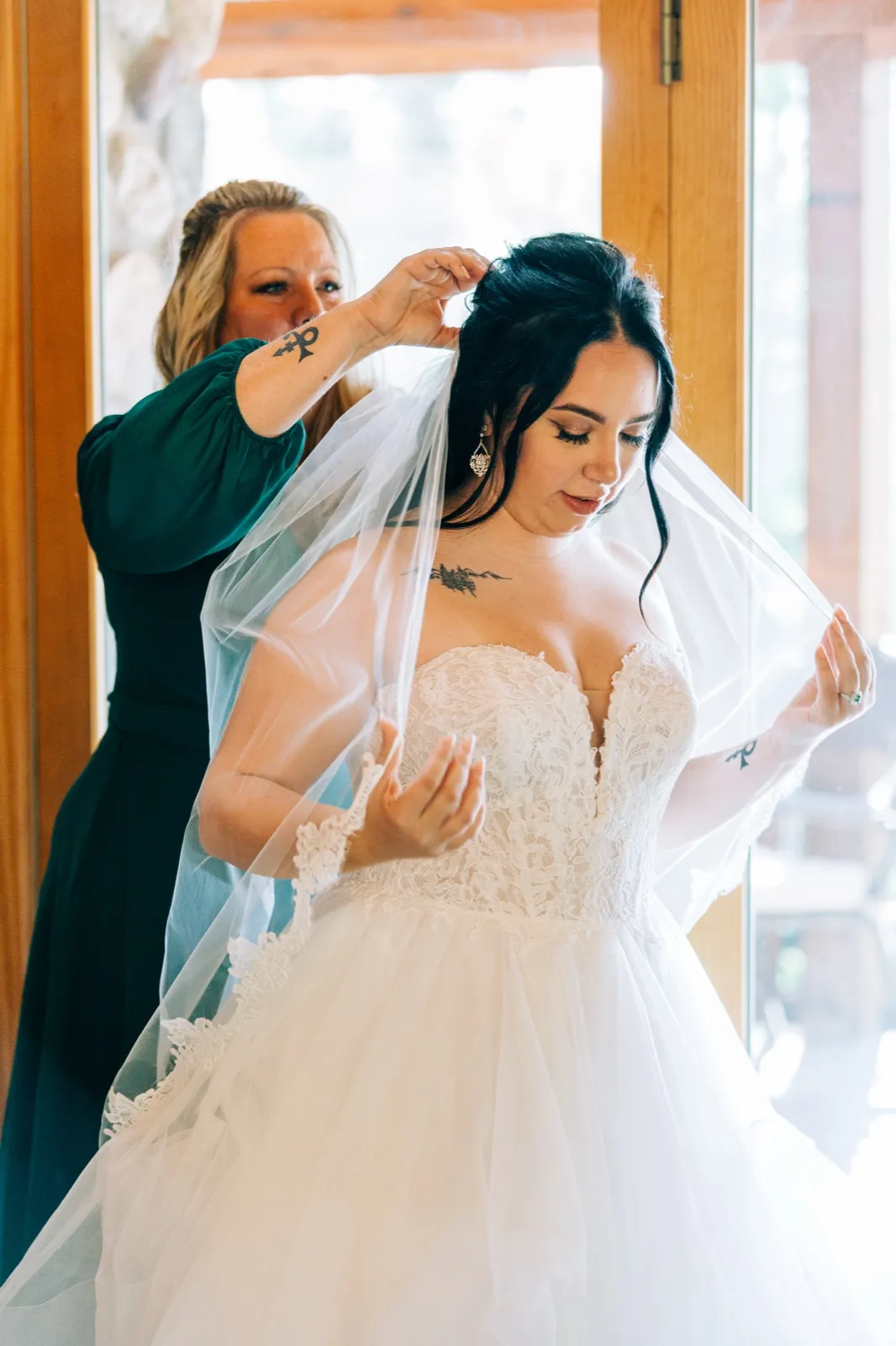 Taylor's mother, in a green dress, lifts her veil from behind as Taylor stands in a strapless lace gown near a sunlit window. — Tim Larsen Photography, Brainerd Lakes MN