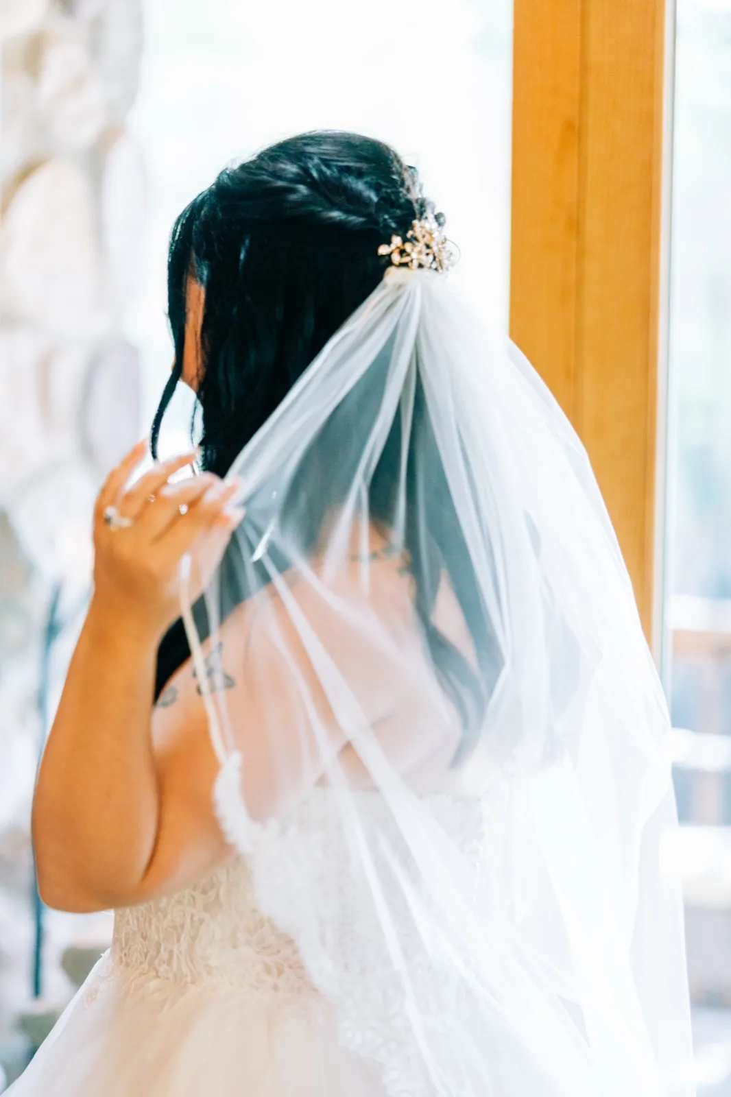 The bride stands at a window with her back to the camera, lifting her translucent veil to reveal a jeweled hair comb — Tim Larsen Photography, Brainerd Lakes MN