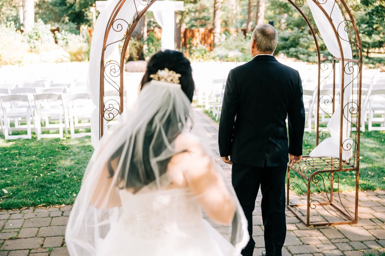The bride walks toward the groom, who waits with his back turned beneath an iron scrollwork arch on a brick aisle in a forest ceremony space — Tim Larsen Photography, Brainerd Lakes MN