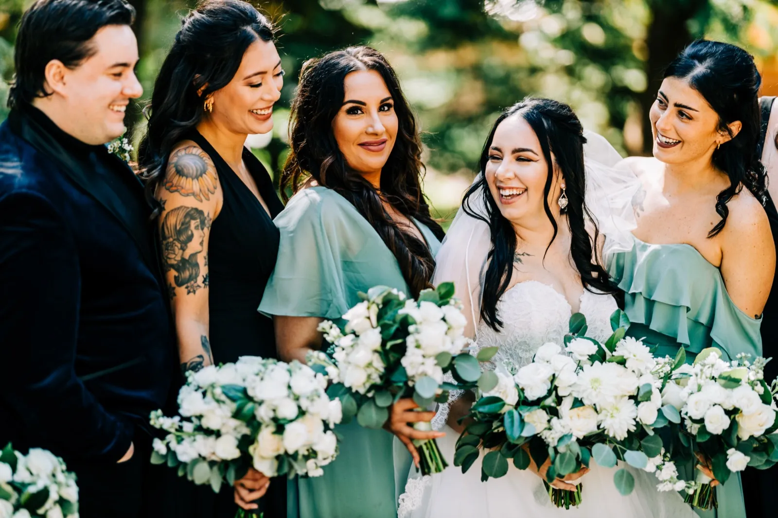 Taylor laughs with three bridesmaids in sage and black dresses and a groomsman in a dark suit, all holding white-and-green bouquets in a sunlit outdoor setting. — Tim Larsen Photography, Brainerd Lakes MN