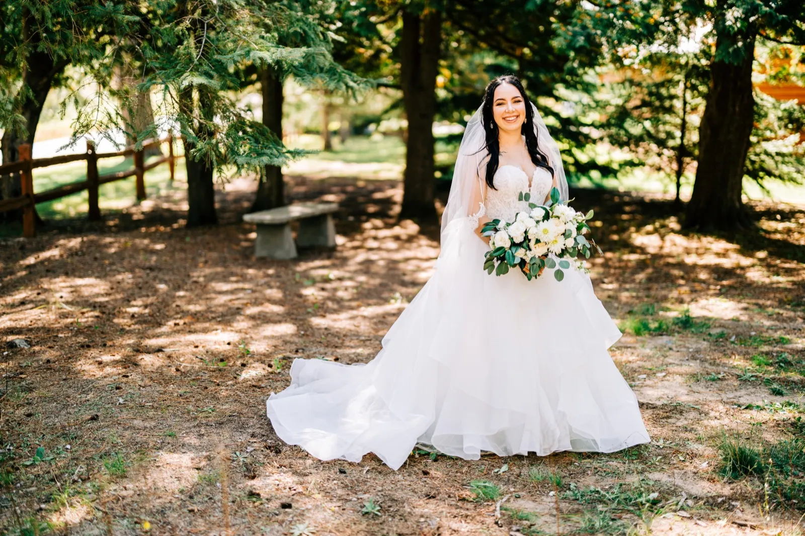 Taylor stands smiling in a full ball gown and long veil, holding a white bouquet, framed by evergreen trees on the wooded grounds of Pine Peaks Event Center. — Tim Larsen Photography, Brainerd Lakes MN
