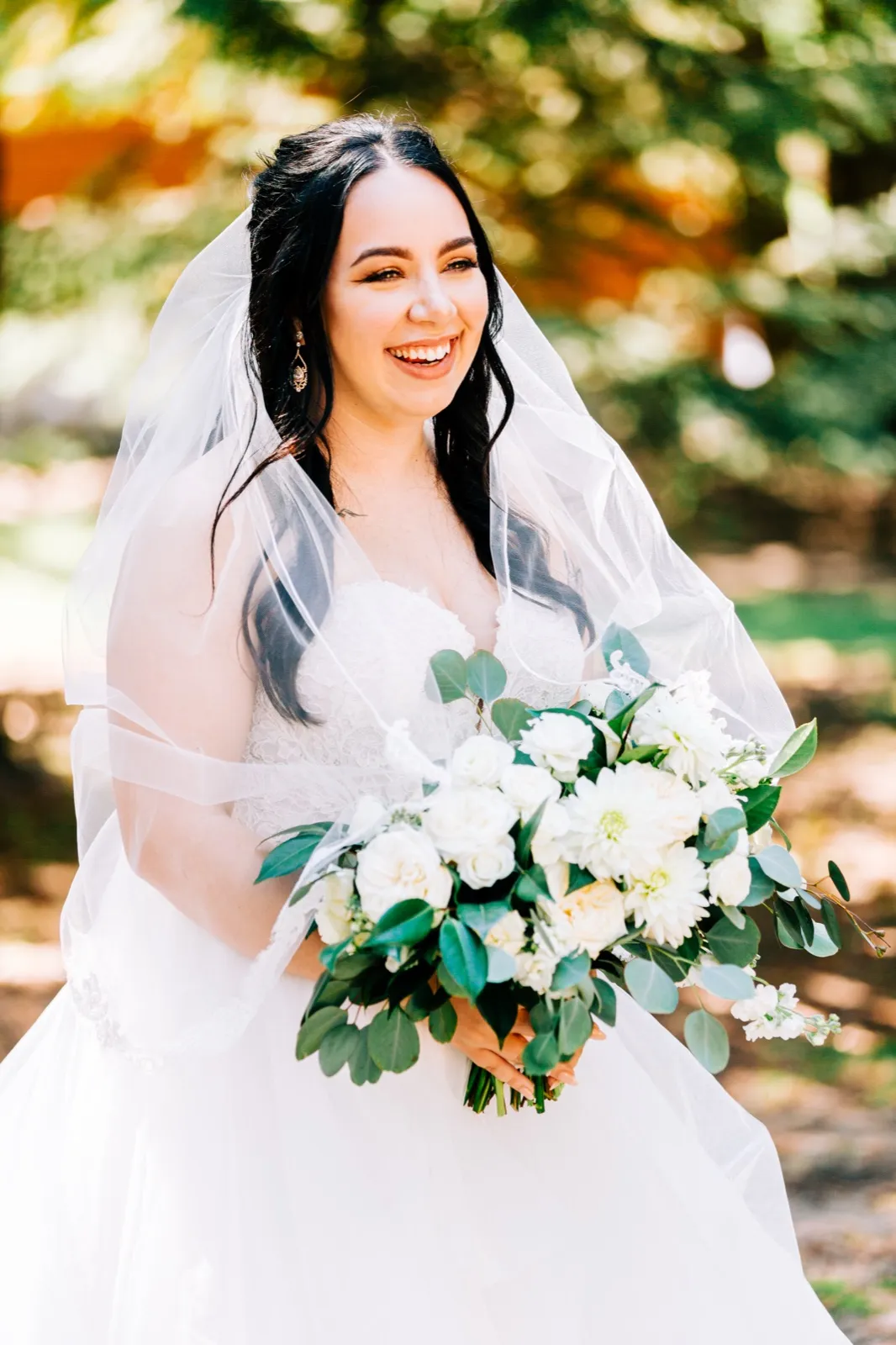 Taylor laughs openly while holding a white and green bouquet, her veil illuminated by sunlight filtering through trees behind her. — Tim Larsen Photography, Brainerd Lakes MN