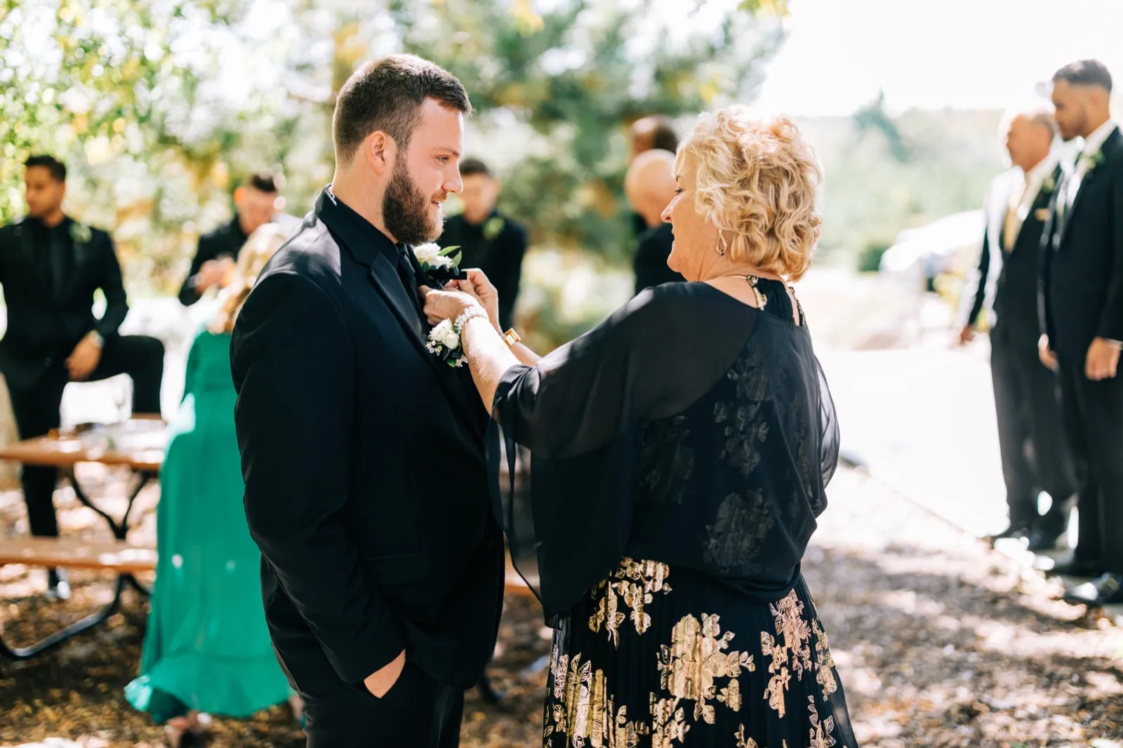 The groom smiles down at his mother as she pins a white boutonniere to his lapel, with groomsmen visible in the sunlit background — Tim Larsen Photography, Brainerd Lakes MN
