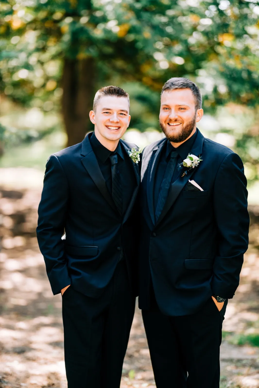 Charles stands beside a groomsman in matching dark suits with white boutonnieres in a sun-dappled wooded clearing. — Tim Larsen Photography, Brainerd Lakes MN