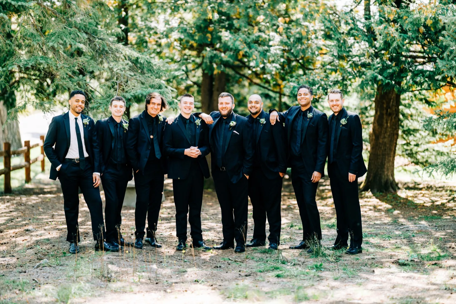 Charles stands at center with seven groomsmen in black suits, arms around each other's shoulders, in a forested clearing at Pine Peaks Event Center. — Tim Larsen Photography, Brainerd Lakes MN