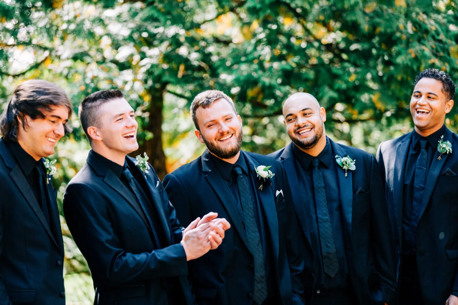 The groom and four groomsmen in black suits laugh together against a backdrop of green forest foliage — Tim Larsen Photography, Brainerd Lakes MN