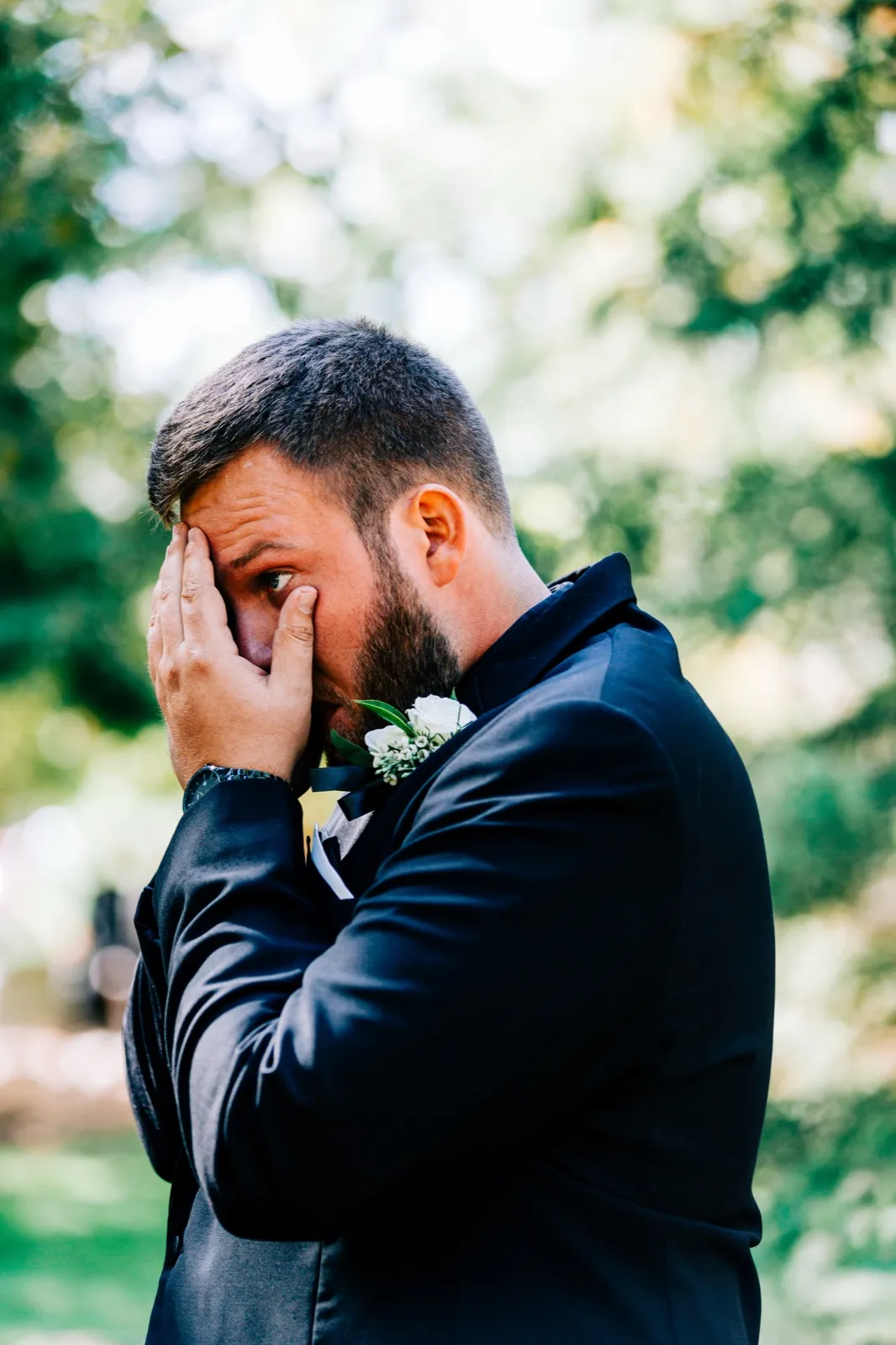 The groom presses a hand to his face, one eye visible between his fingers, as he cries during the first look in a wooded clearing — Tim Larsen Photography, Brainerd Lakes MN
