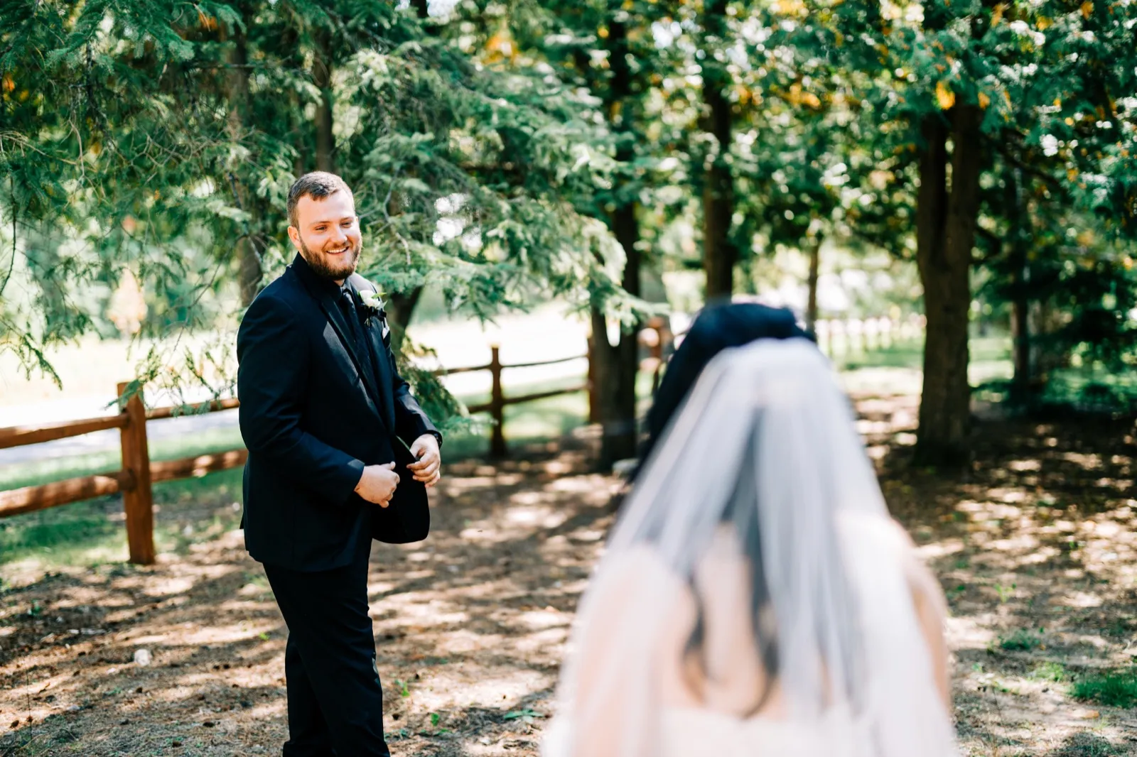 The groom smiles broadly as he sees the bride for the first time, her veil out of focus in the foreground along a wooded path — Tim Larsen Photography, Brainerd Lakes MN