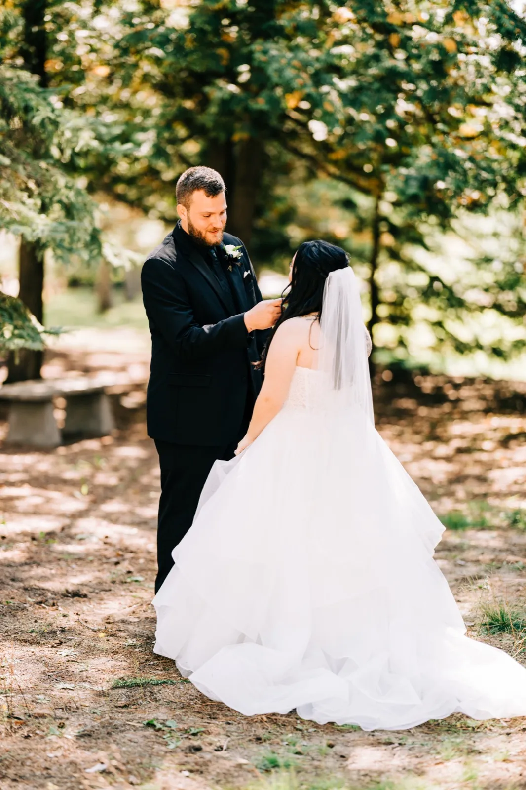 Charles adjusts something on Taylor's bodice as she stands with her back to the camera, full veil and trained gown spread across a sun-dappled forest clearing. — Tim Larsen Photography, Brainerd Lakes MN