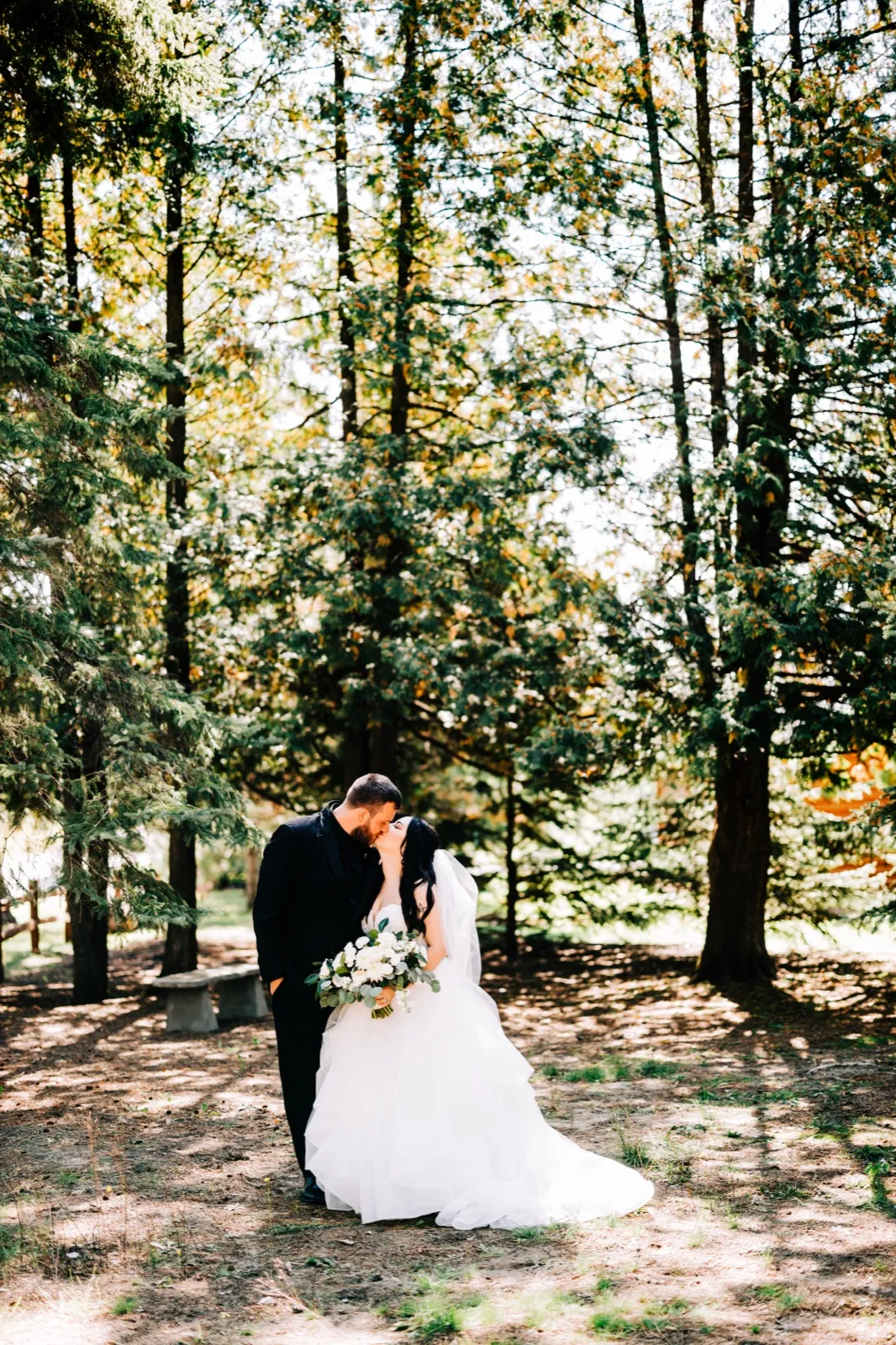 Taylor and Charles share a kiss in a clearing surrounded by towering evergreen pines, Taylor's veil and full gown trailing behind her as she holds a white and green bouquet. — Tim Larsen Photography, Brainerd Lakes MN