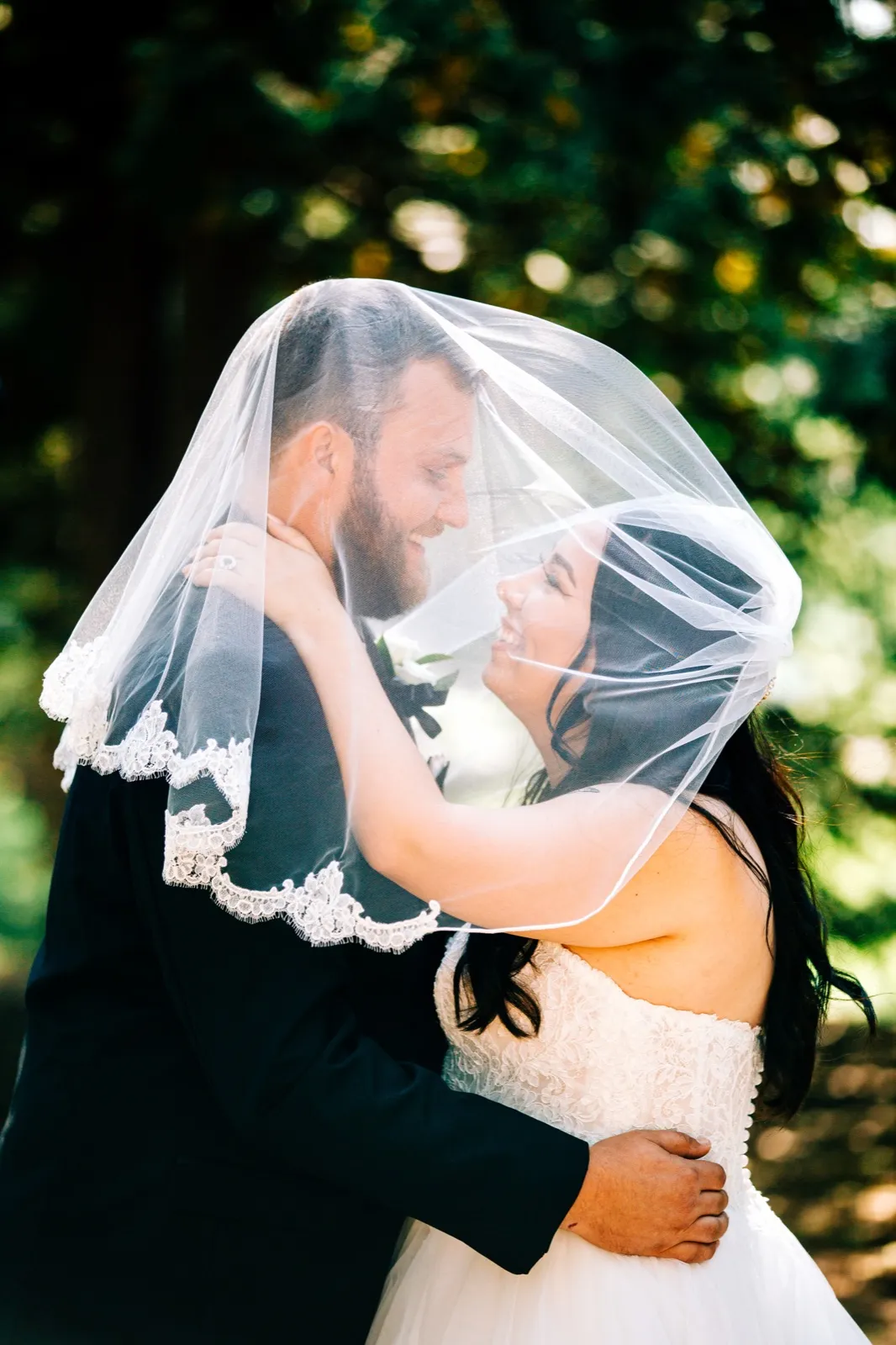Taylor and Charles laugh face-to-face beneath her sunlit veil, framed by soft green forest light. — Tim Larsen Photography, Brainerd Lakes MN