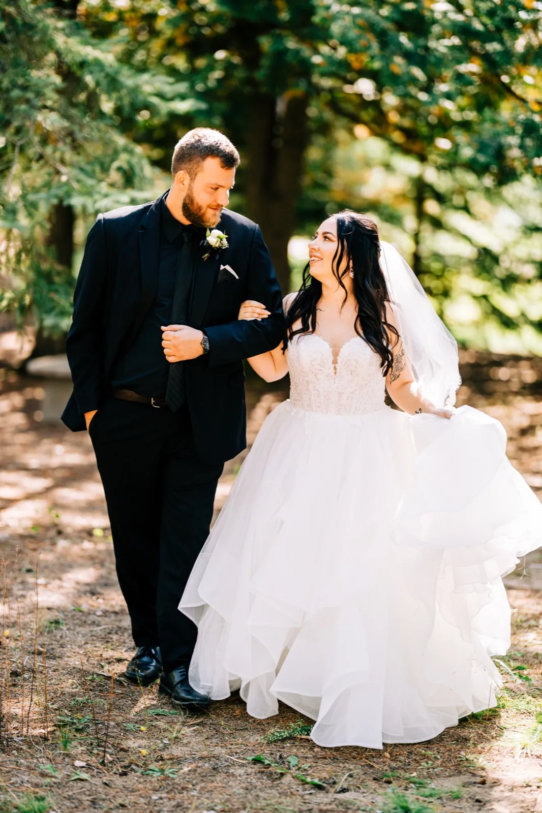 Taylor lifts the layered tulle of her gown as she walks through a sunlit forest path, looking up at Charles in a black suit beside her. — Tim Larsen Photography, Brainerd Lakes MN