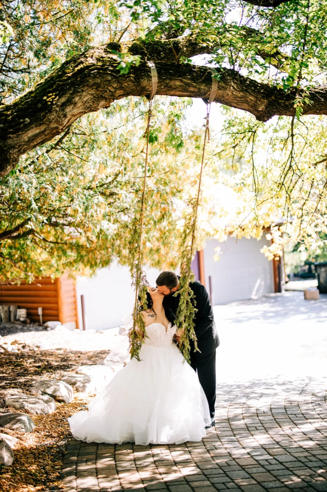 Taylor and Charles kiss while seated on a floral-wrapped swing hanging from a sprawling tree branch on a paved courtyard at Pine Peaks Event Center. — Tim Larsen Photography, Brainerd Lakes MN
